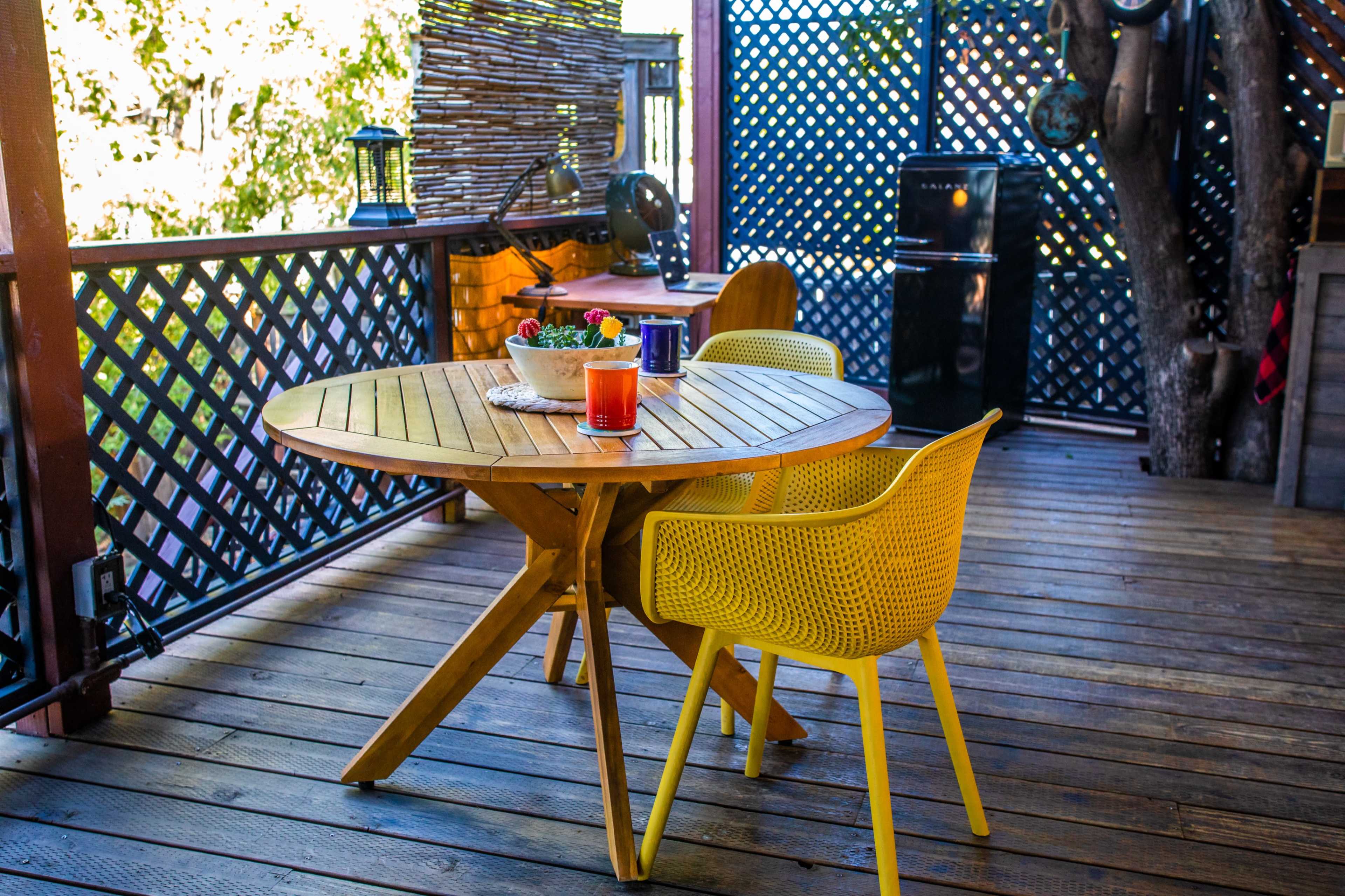 A wooden table with two yellow chairs is set on a wooden deck, surrounded by a patterned lattice and outdoor decor.