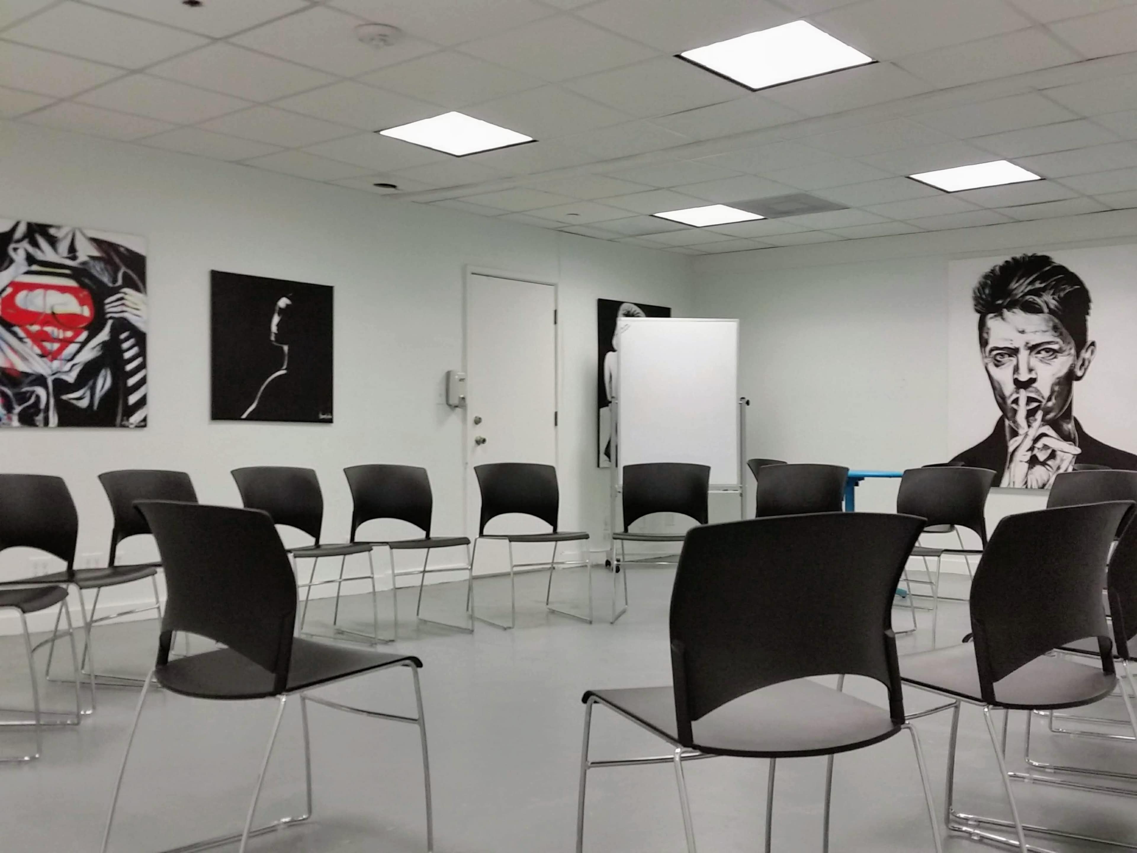 A meeting room with black chairs arranged in a circle, featuring artwork on the walls, including a superhero symbol and a portrait of a man.