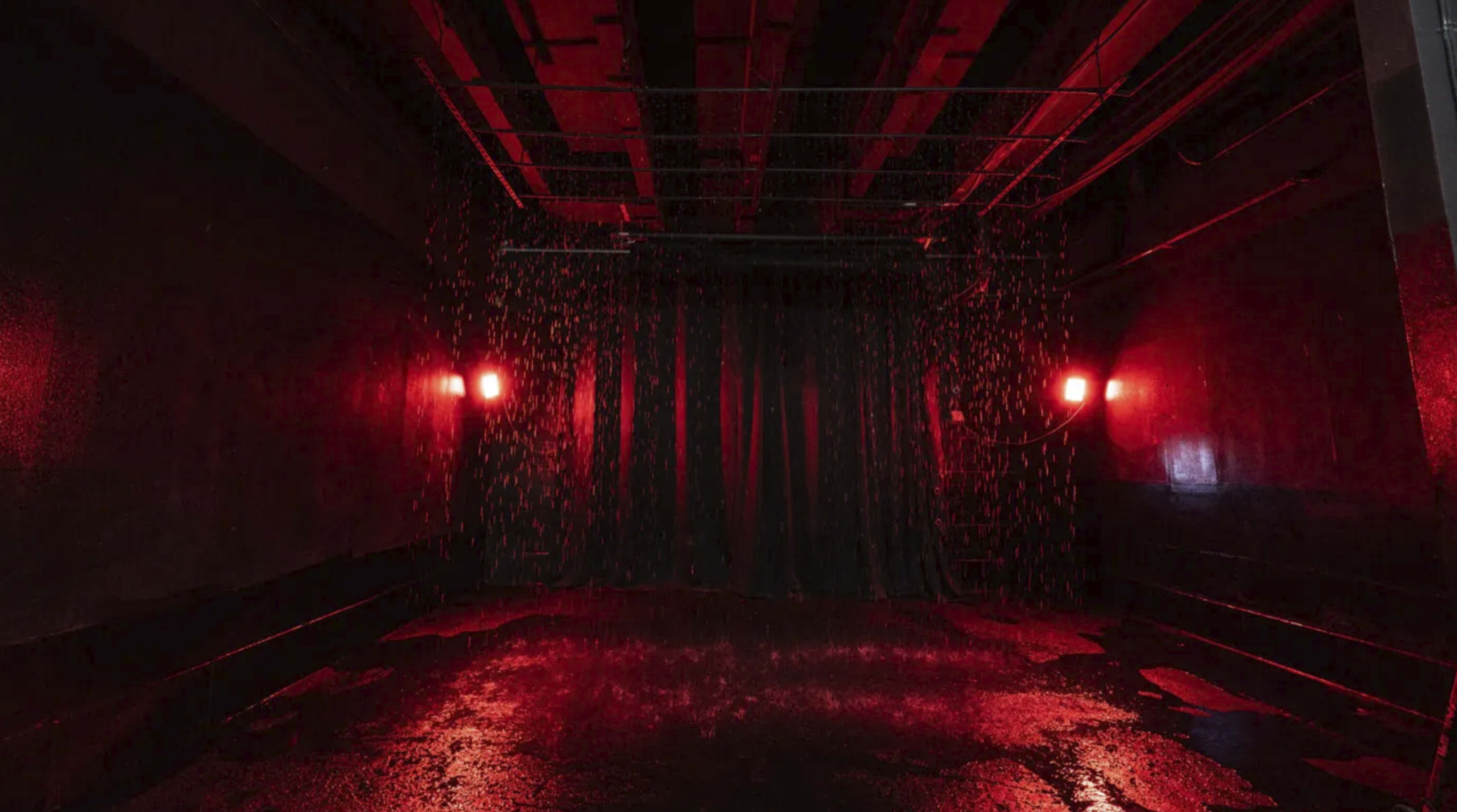 A dark room with red lighting, a black curtain in the background, and water cascading down from the ceiling onto the wet floor.