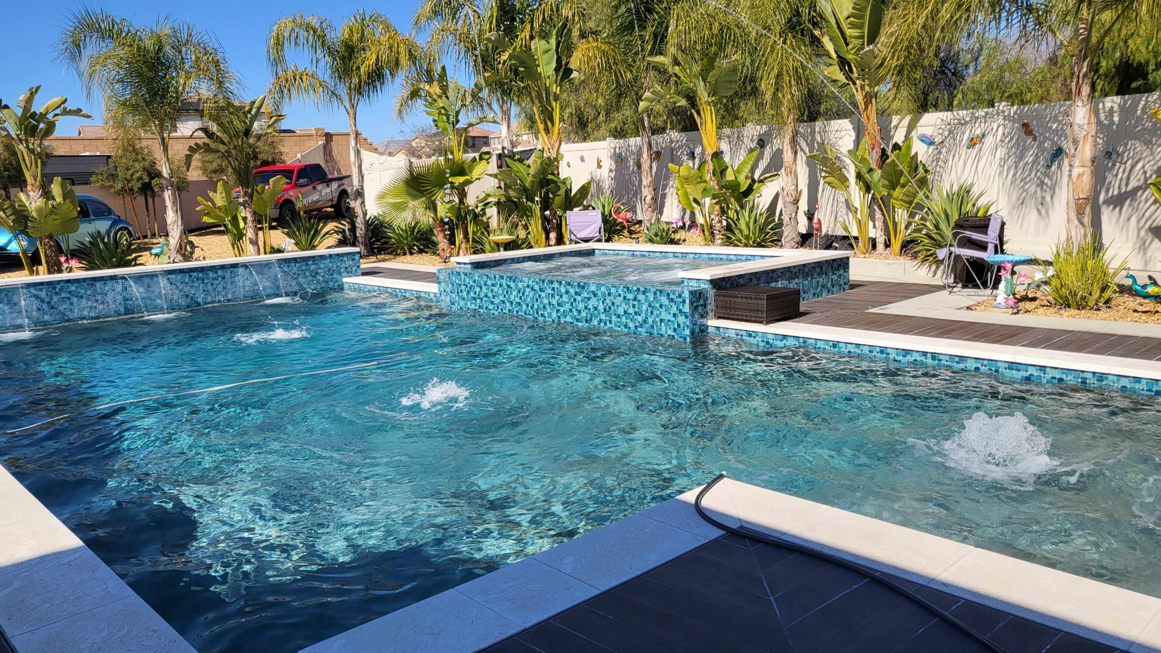 A swimming pool area featuring a main pool with a spa section, surrounded by palm trees and landscaped plants.