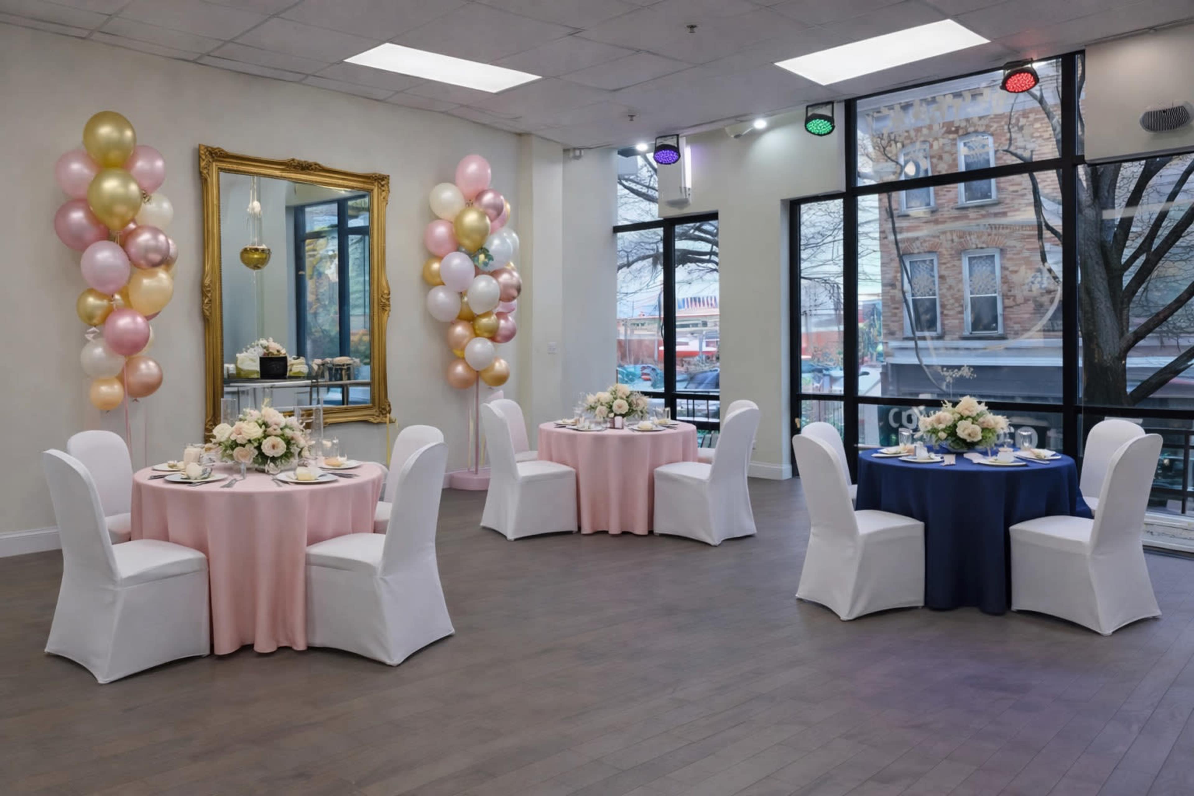 The image shows a decorated event space with round tables draped in pink and blue tablecloths, each set with floral centerpieces and surrounded by white chairs, while colorful balloons are positioned at the corners and large windows offer a view of the outside.