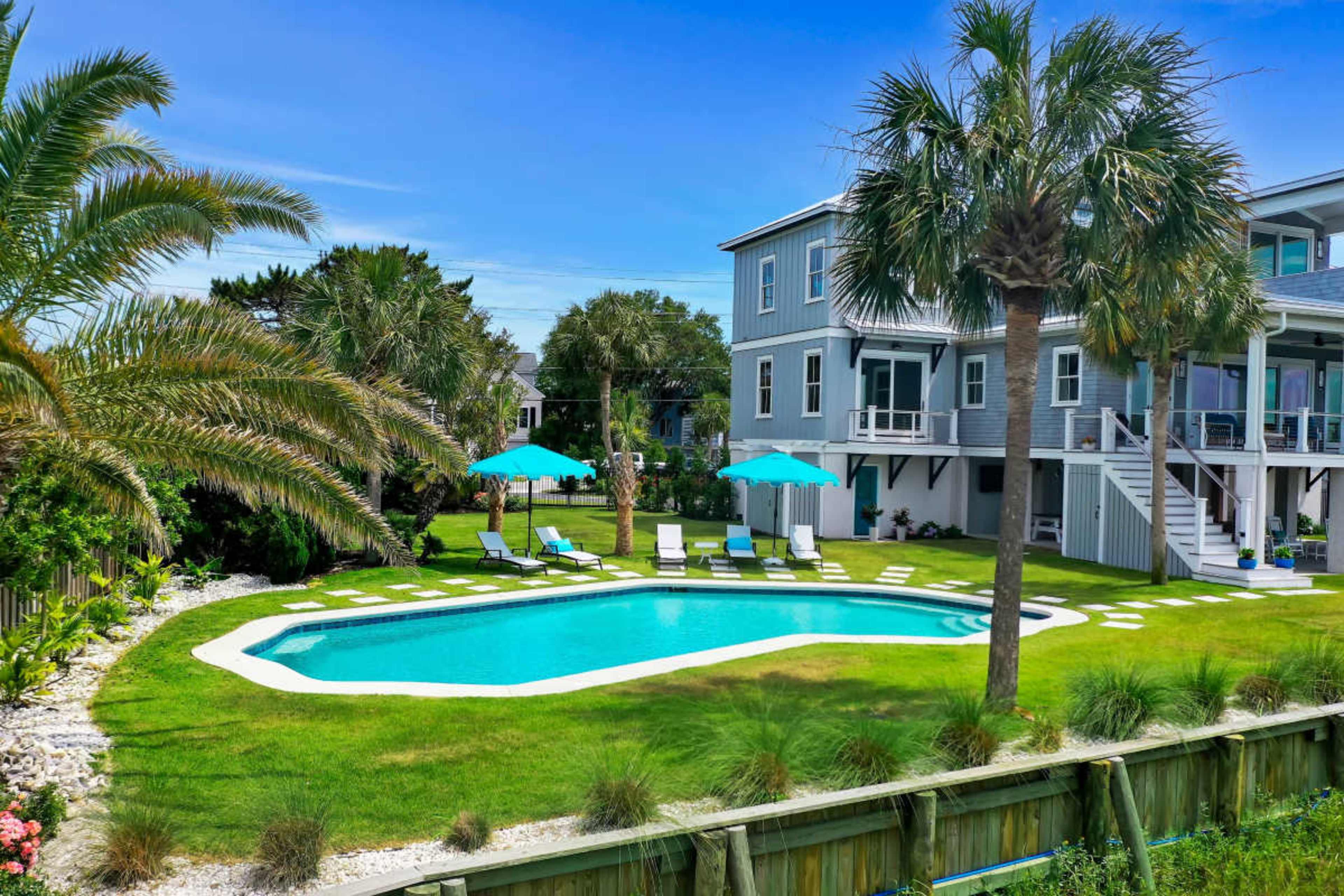 The image shows a private swimming pool surrounded by palm trees and lounge chairs, with a two-story house in the background.