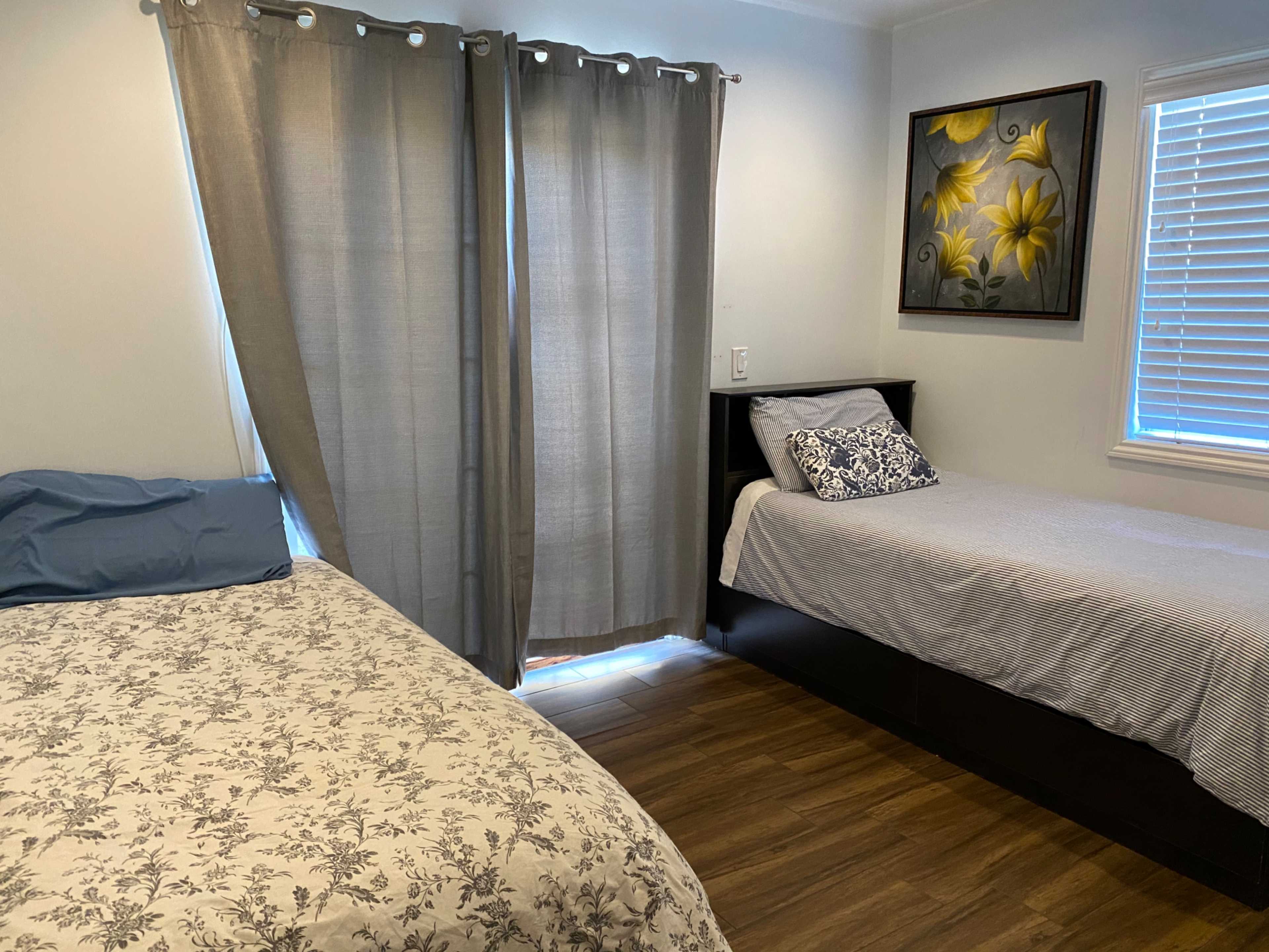 A bedroom with a double bed covered in a floral comforter beside a twin bed against a wall, with gray curtains and a painting of yellow flowers on the wall.