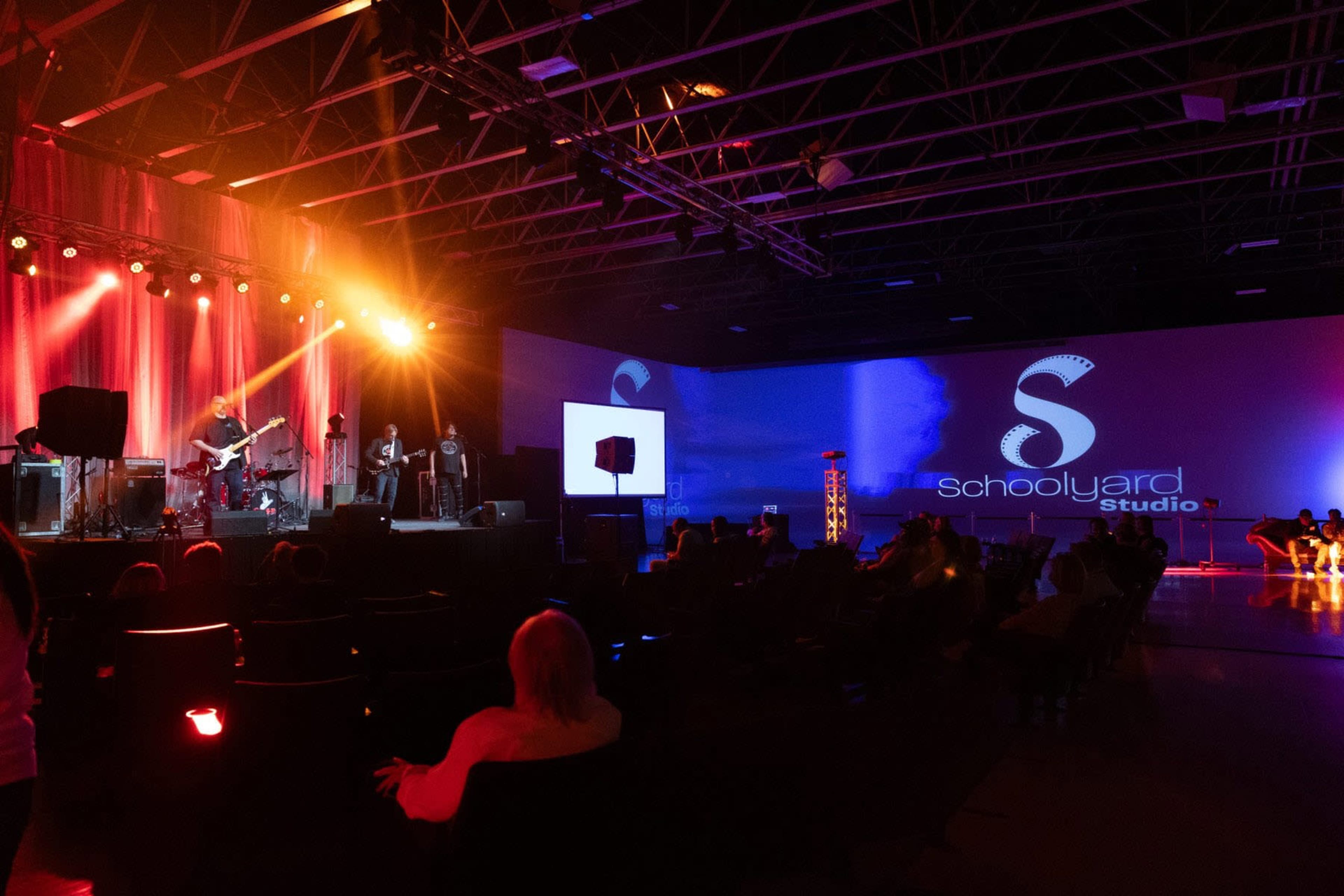 A live music performance is taking place on a stage, illuminated by colorful lights, while an audience sits in rows of chairs.