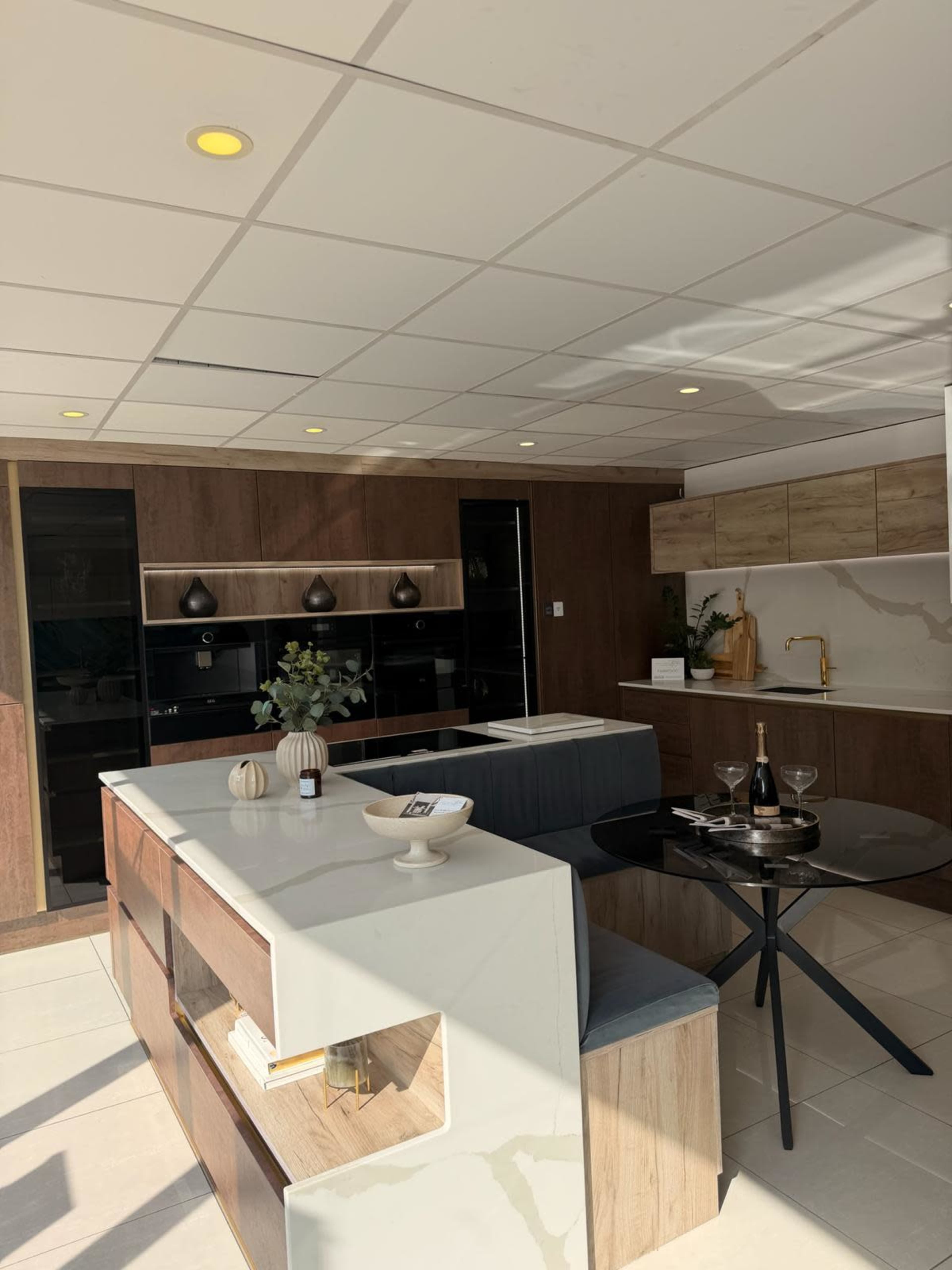The image shows a modern kitchen featuring wooden cabinetry, a central island with seating, and a dining area with a round black table.