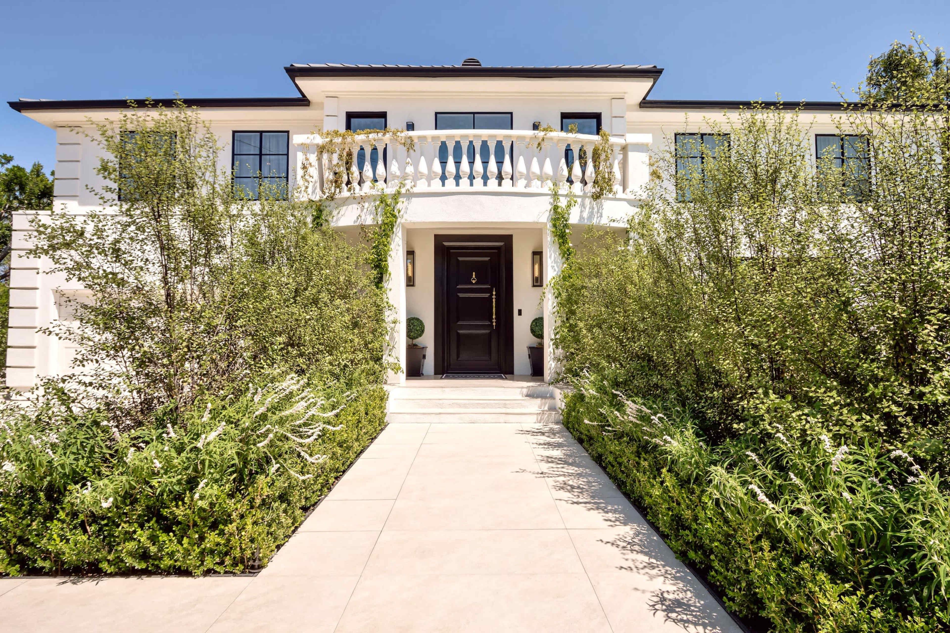 The image shows a modern two-story house with a symmetrical façade, a front balcony, and landscaped greenery surrounding the entrance.
