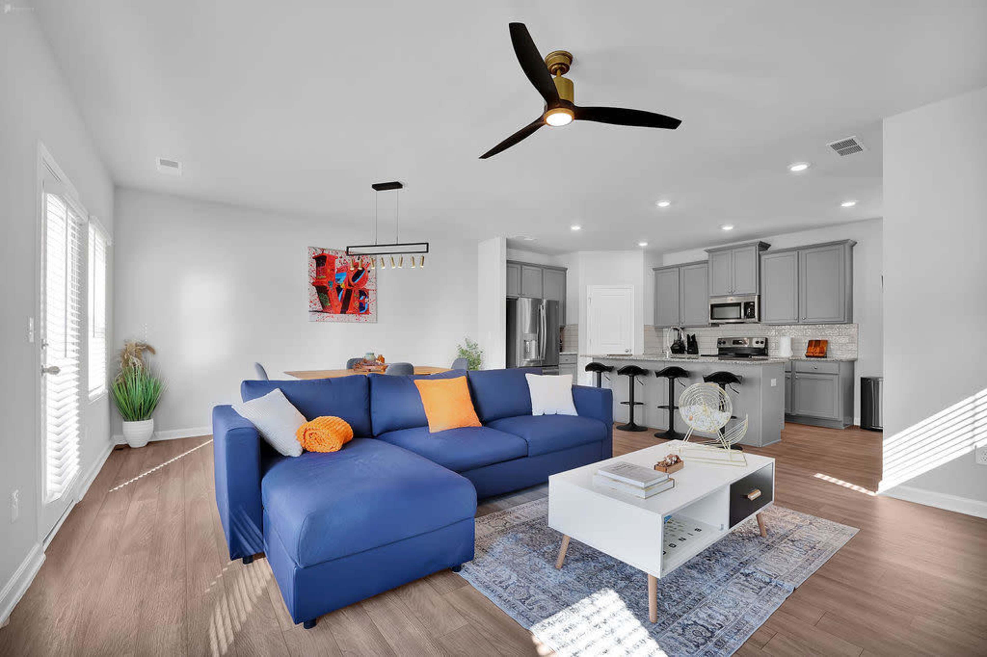 A modern living room with a blue sectional sofa, a coffee table, and a kitchen area in the background featuring gray cabinets and stainless steel appliances.