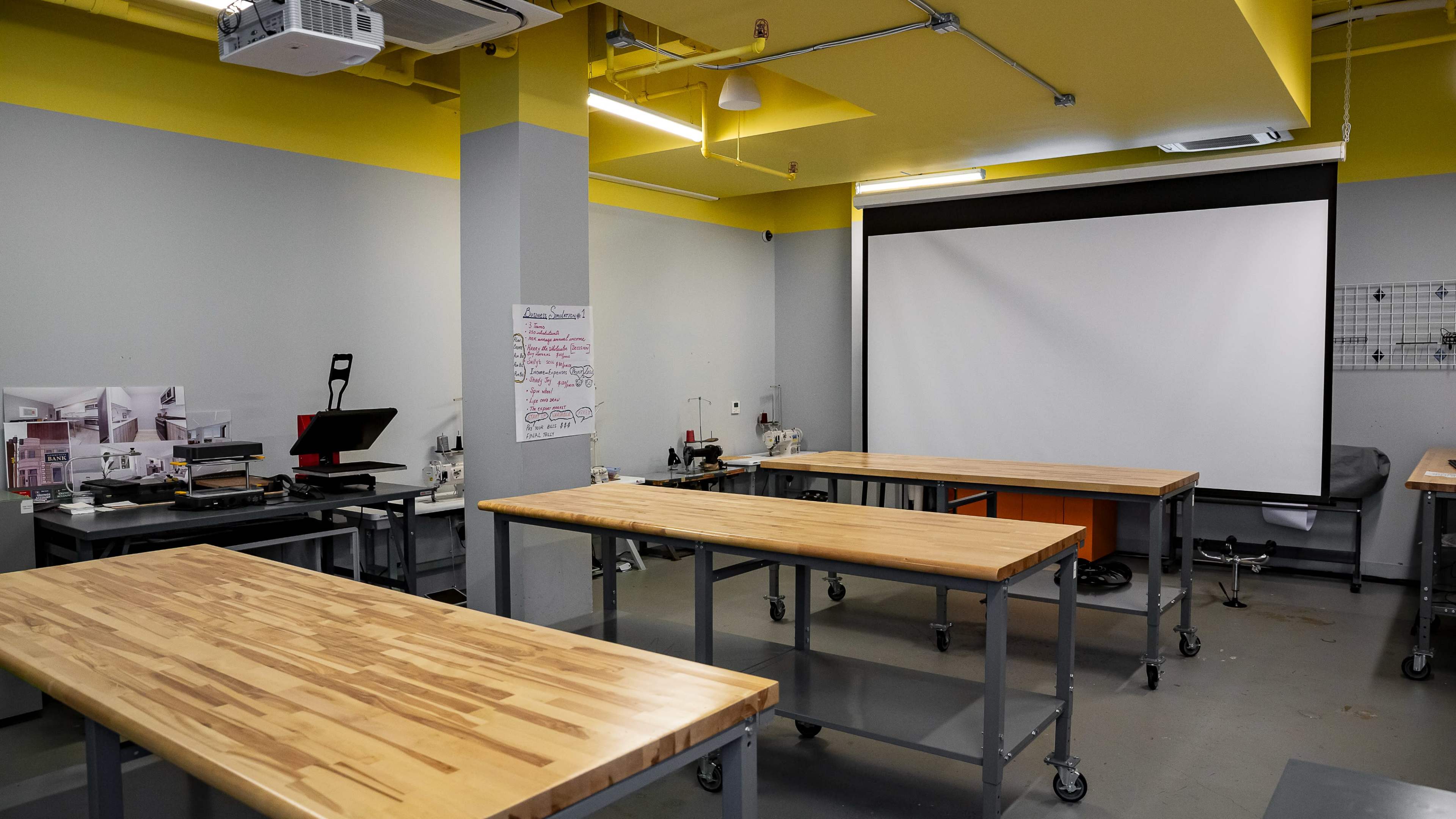 A spacious workshop is equipped with wooden tables, a projector screen, and various tools on counters and shelves.
