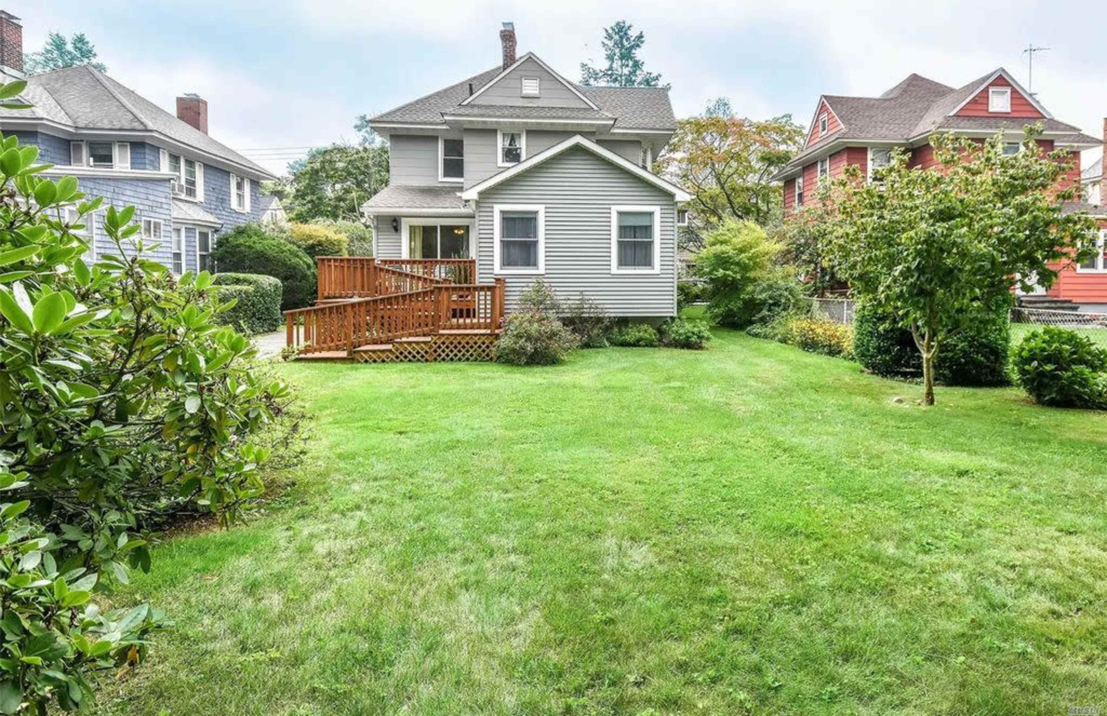 A residential yard with a two-story house, a wooden deck, and well-maintained grass surrounded by shrubs and trees.