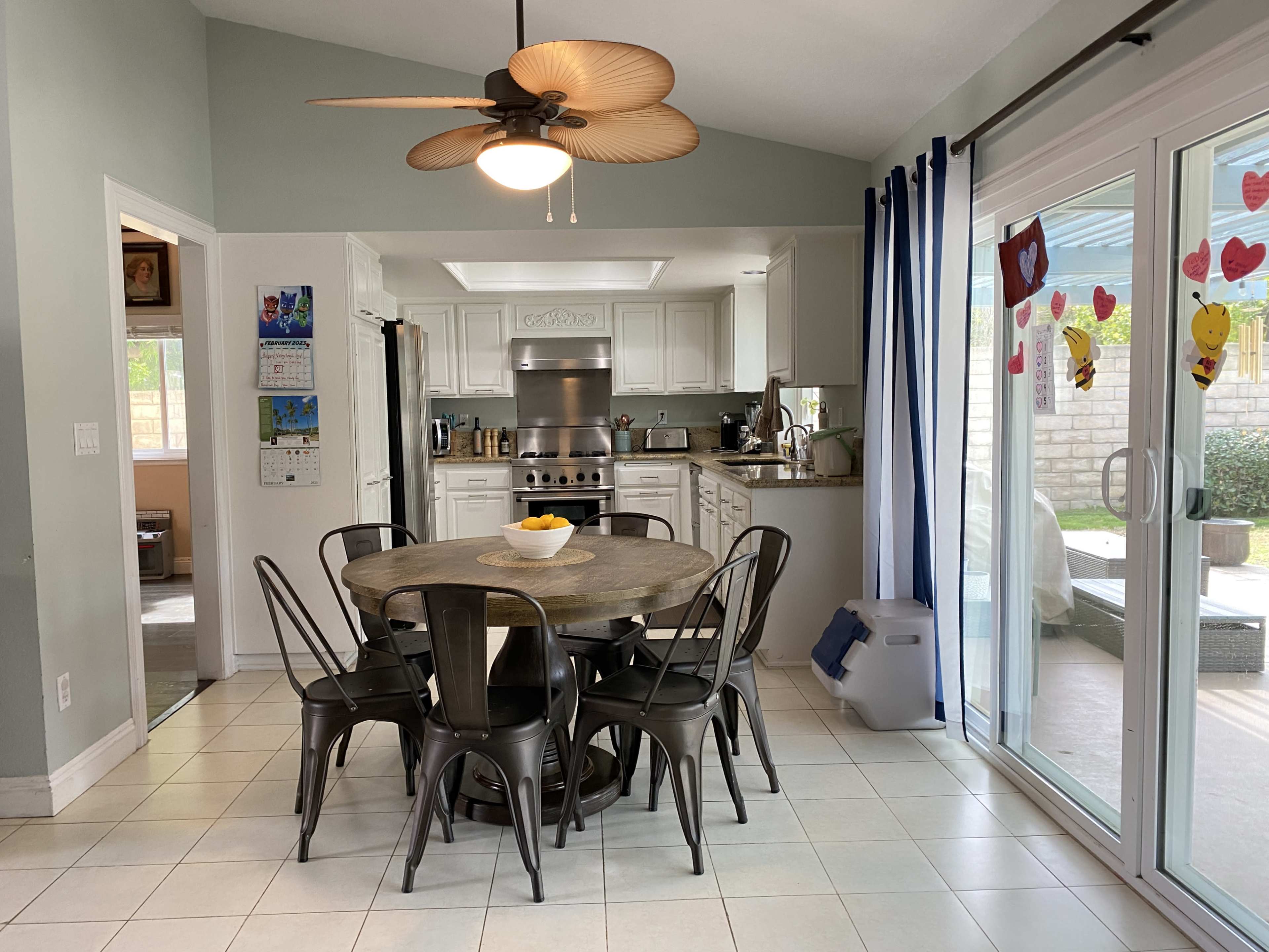 A modern kitchen and dining area feature a round wooden table surrounded by metal chairs, with a ceiling fan overhead and large glass doors leading to an outdoor space.