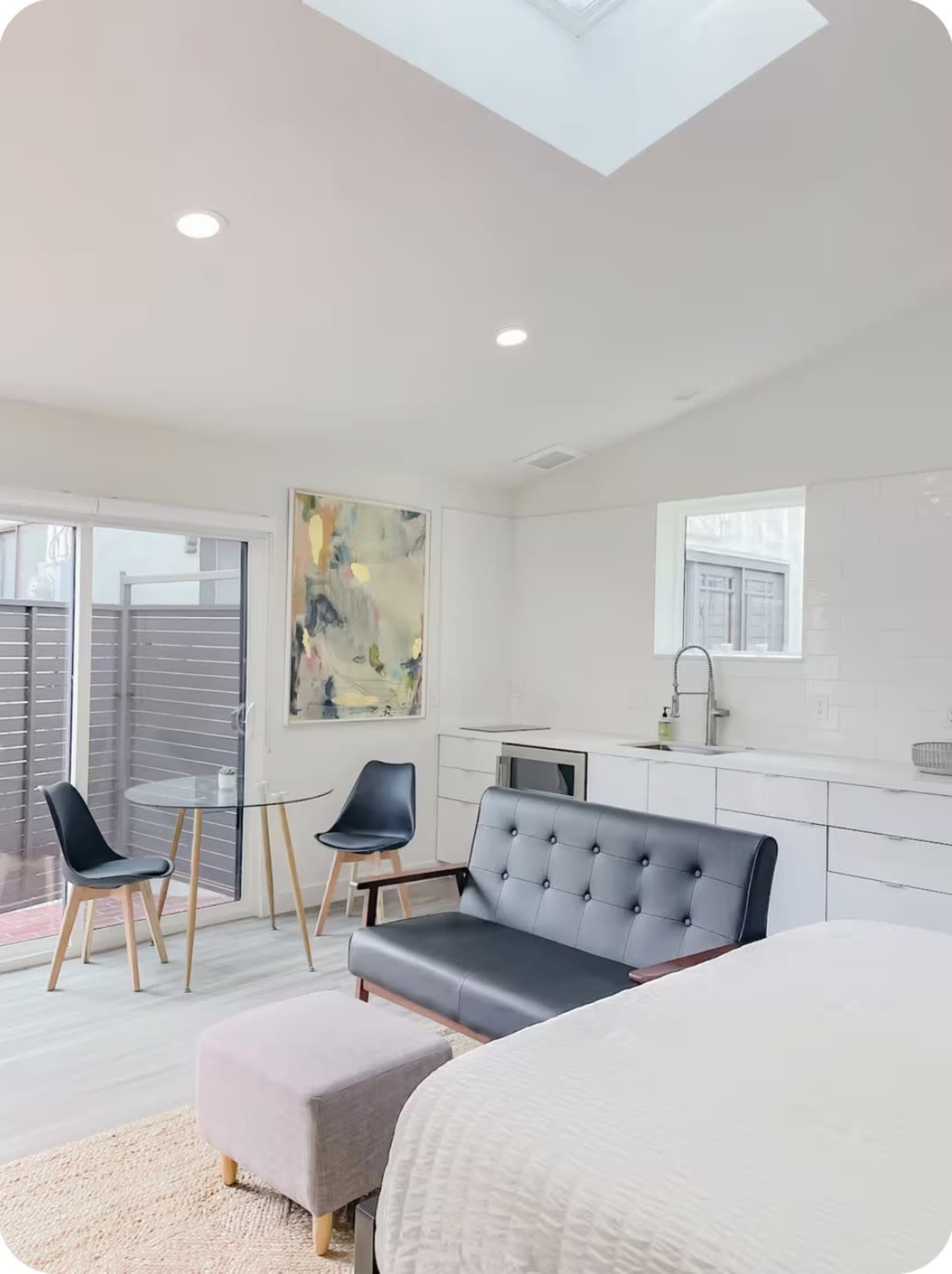 A modern, open-concept living space features a black sofa, a small dining table, and a skylight, with white walls and wooden accents.