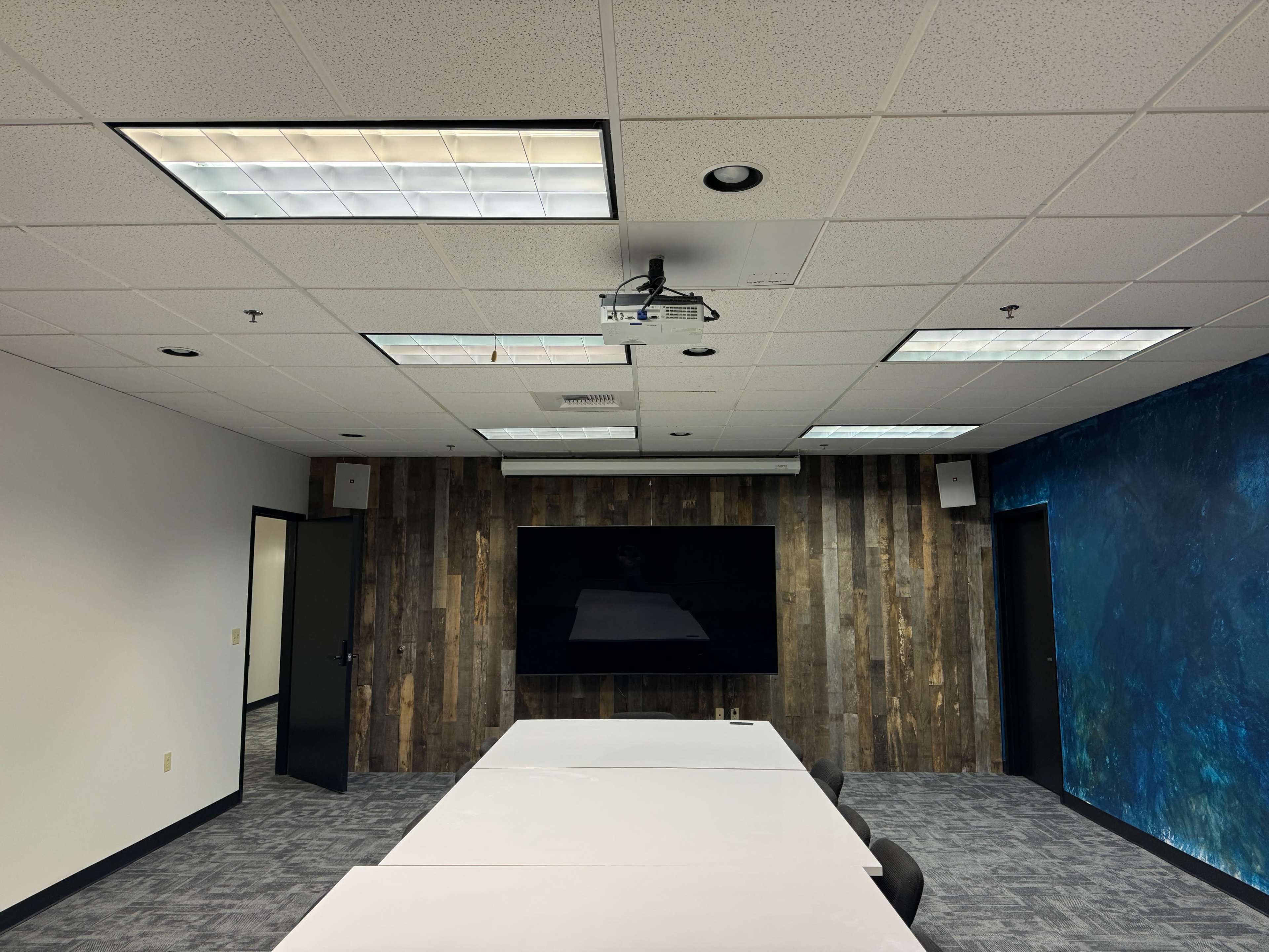 A modern meeting room features a long white table, a large wall-mounted screen, and a textured wooden accent wall, with overhead lights and a projector on the ceiling.