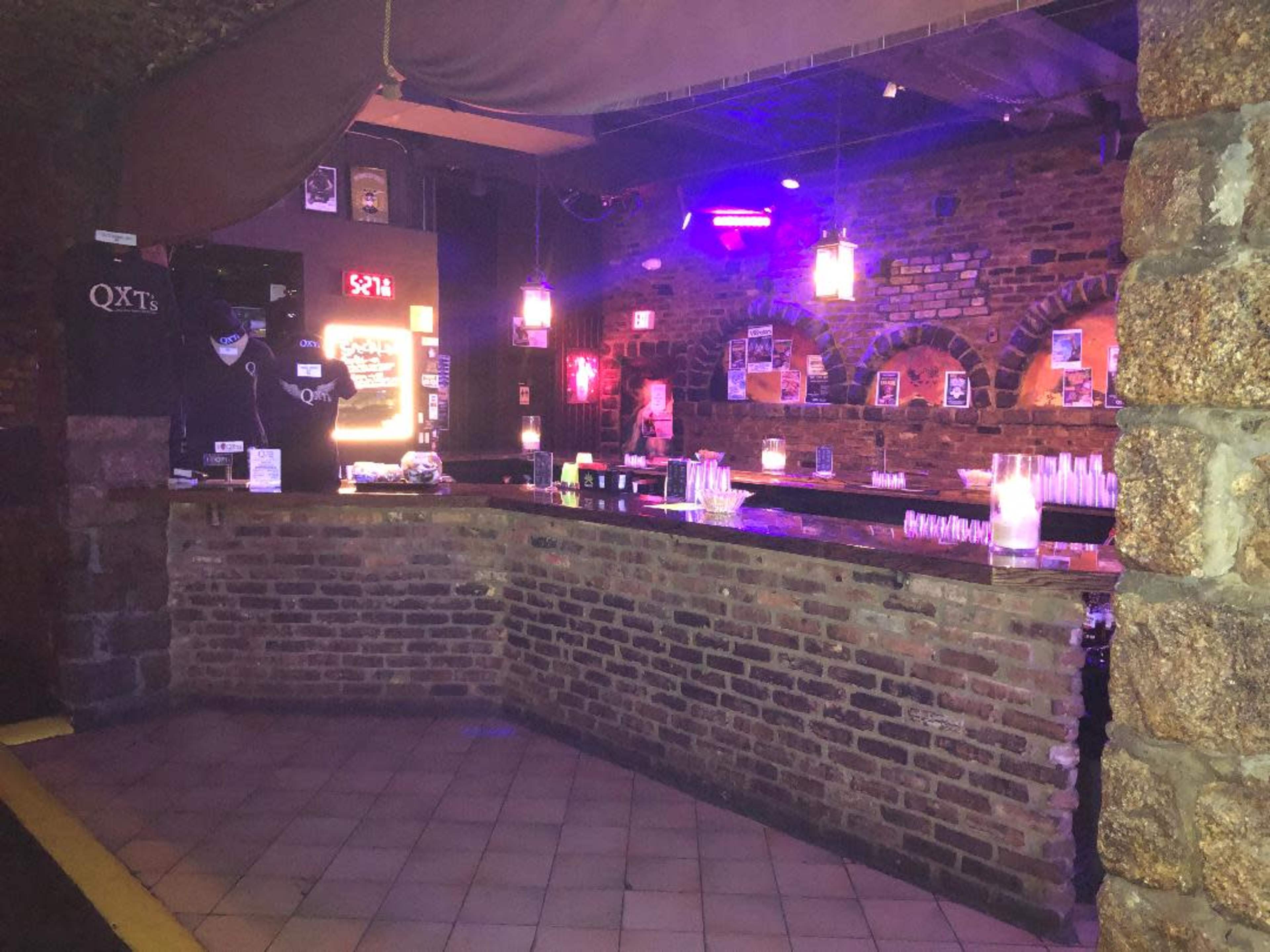 A dimly lit bar features a brick wall backdrop and a long counter with illuminated glassware and hanging light fixtures.