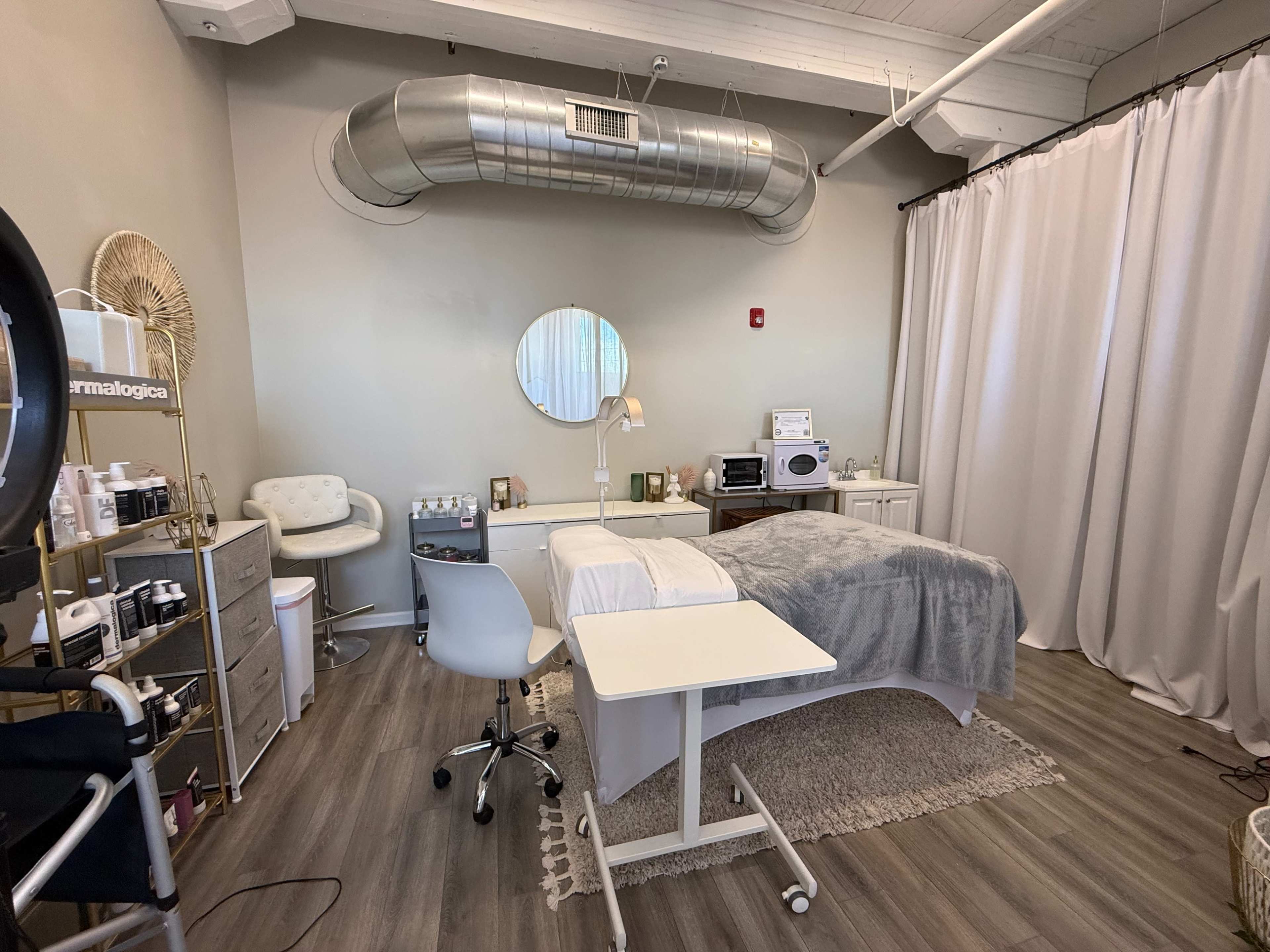 The image shows a well-organized beauty treatment room with a bed, a chair, and various skincare products displayed on shelves.