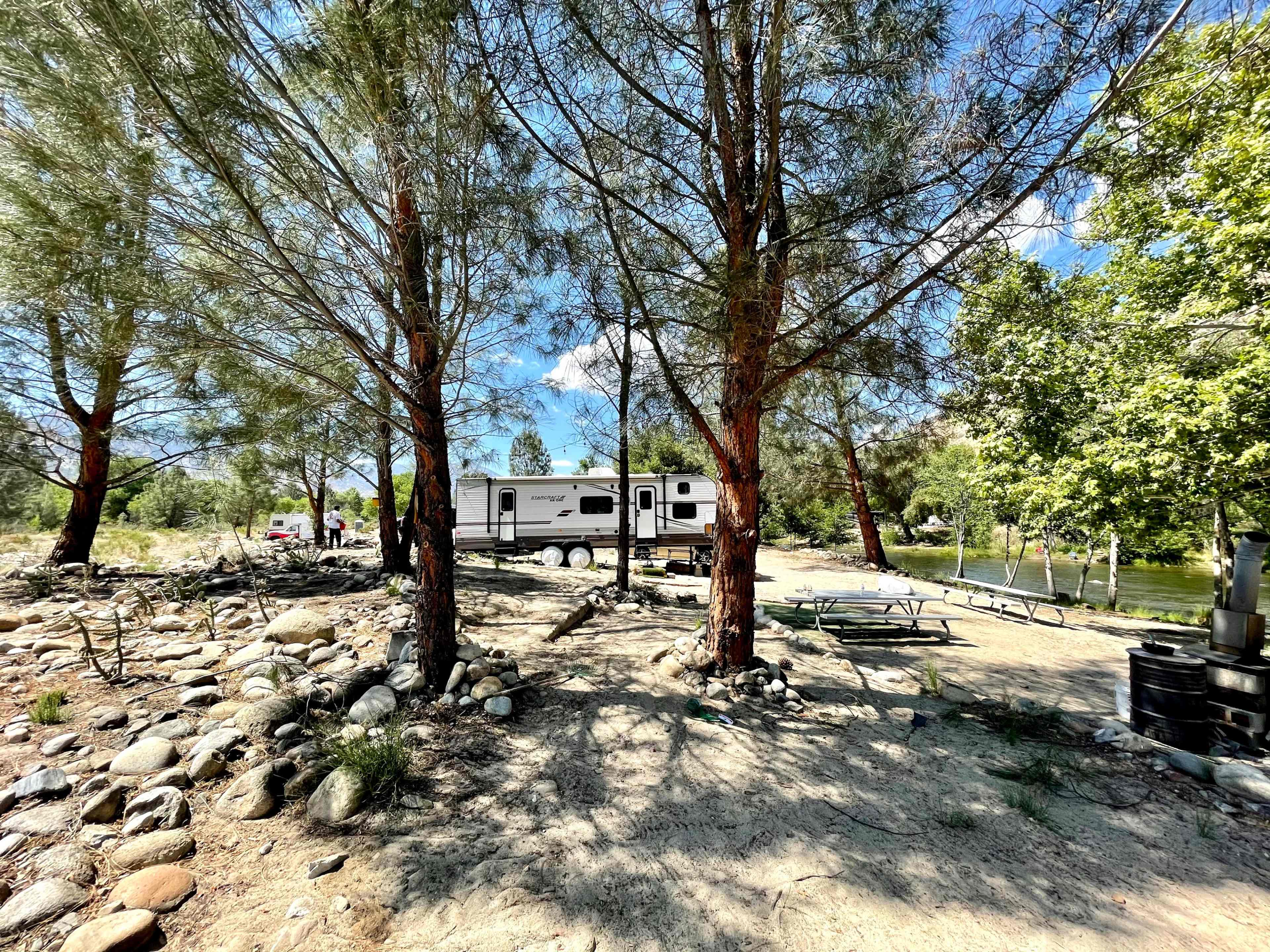 A recreational vehicle is parked among trees near a river, with a picnic area and boulders in the foreground.