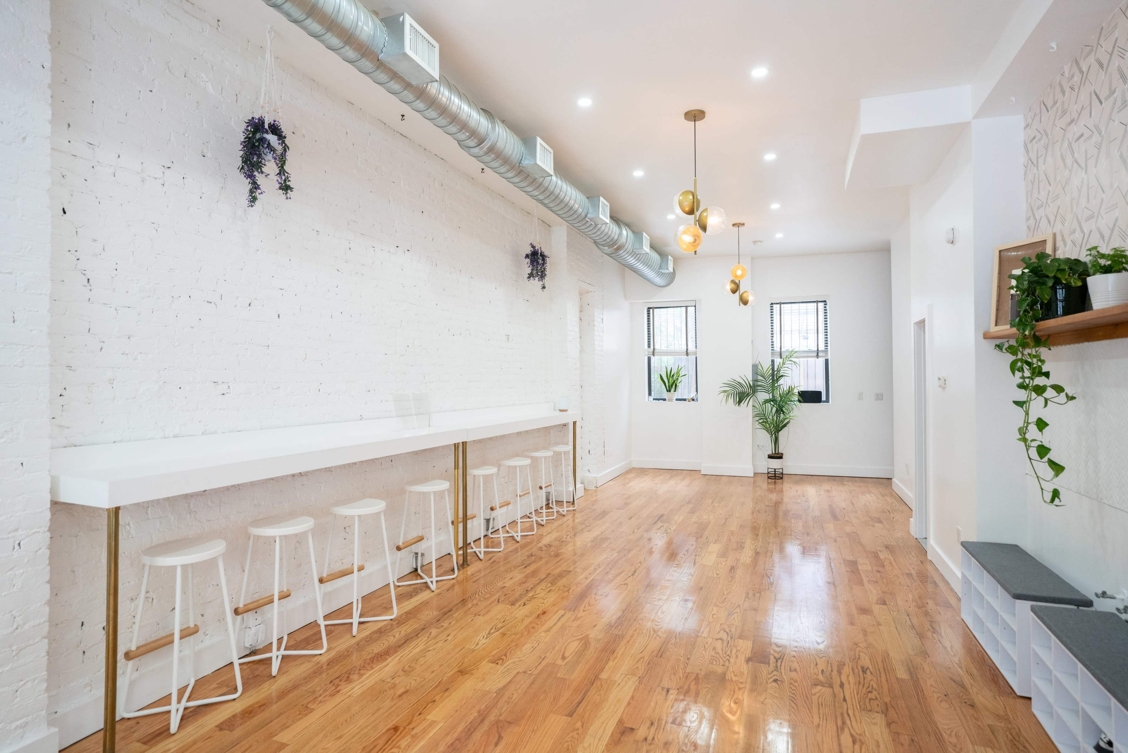 A spacious interior with white walls, wooden floors, a long bar area with white stools, and several plants.