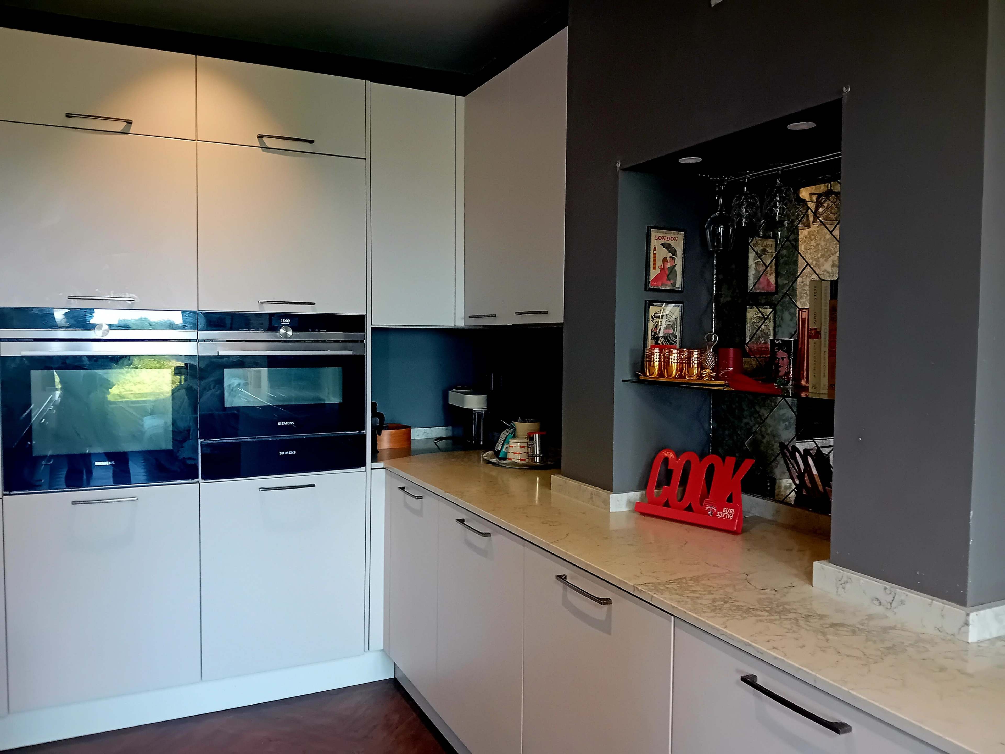 The image shows a modern kitchen with sleek white cabinetry, built-in ovens, and a countertop featuring a decorative "COOK" sign.