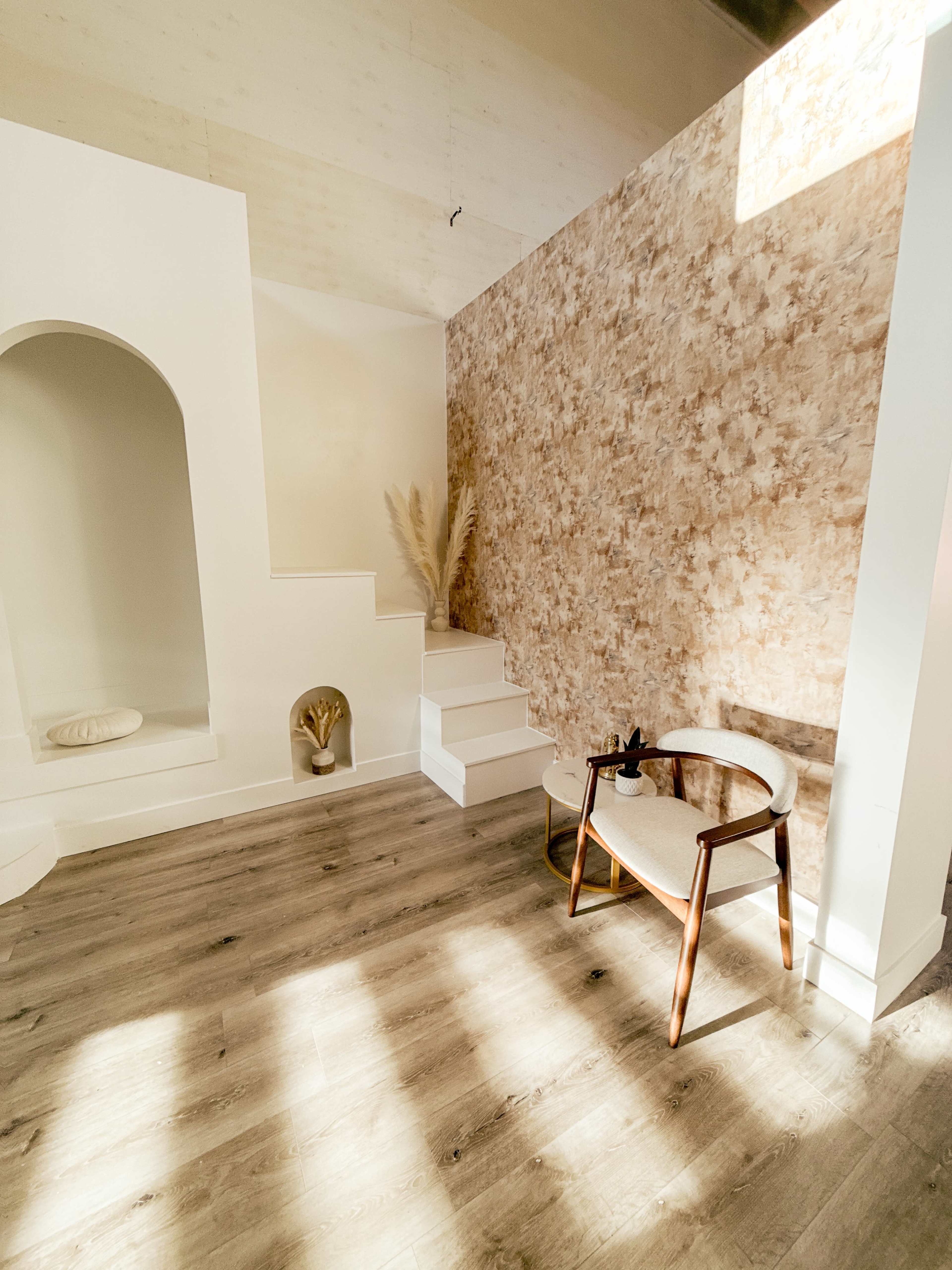 The image shows a well-lit interior space featuring a staircase, a decorative wall with a floral pattern, and a modern wooden chair.