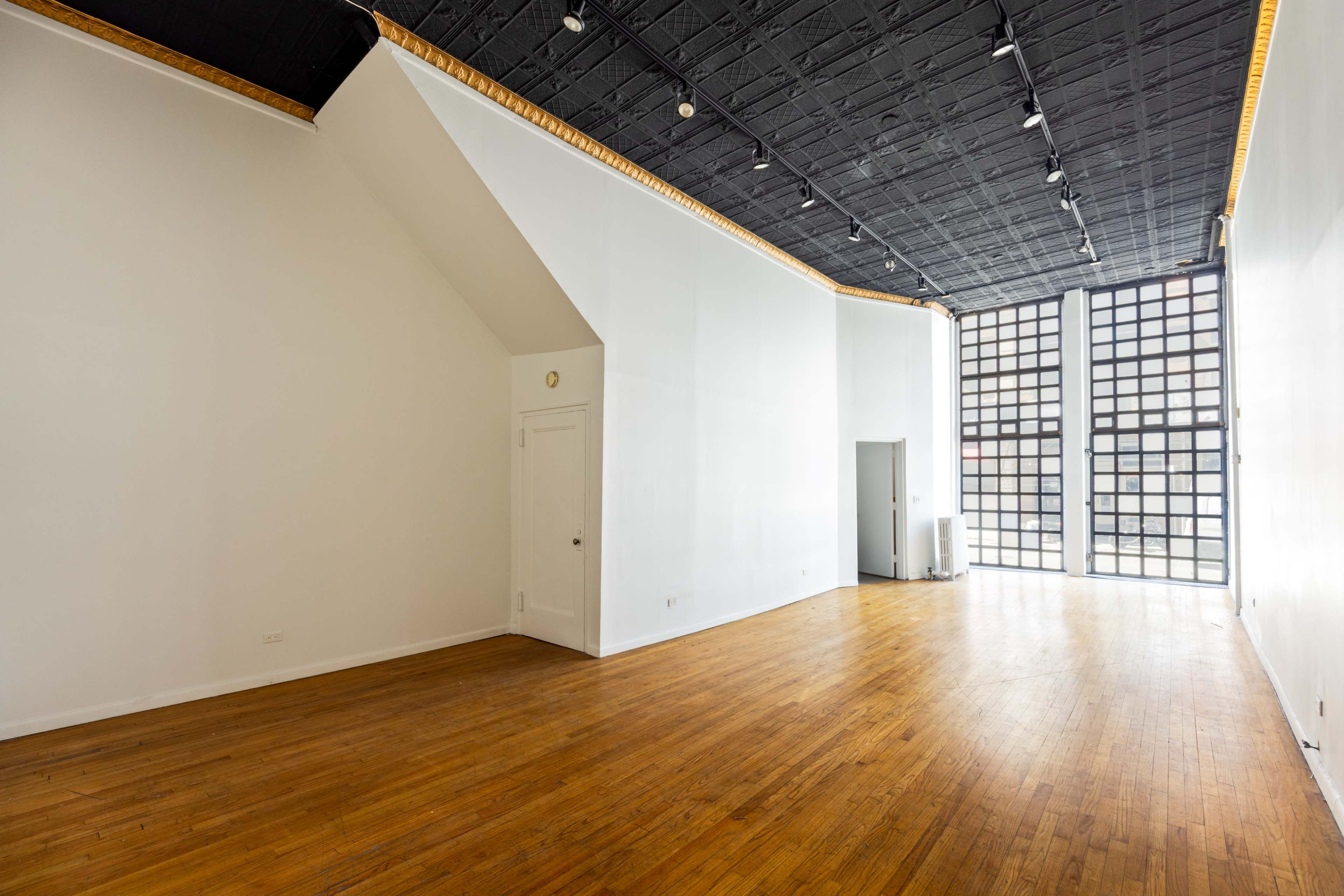The image shows a spacious, empty room with hardwood floors and large windows on one wall.