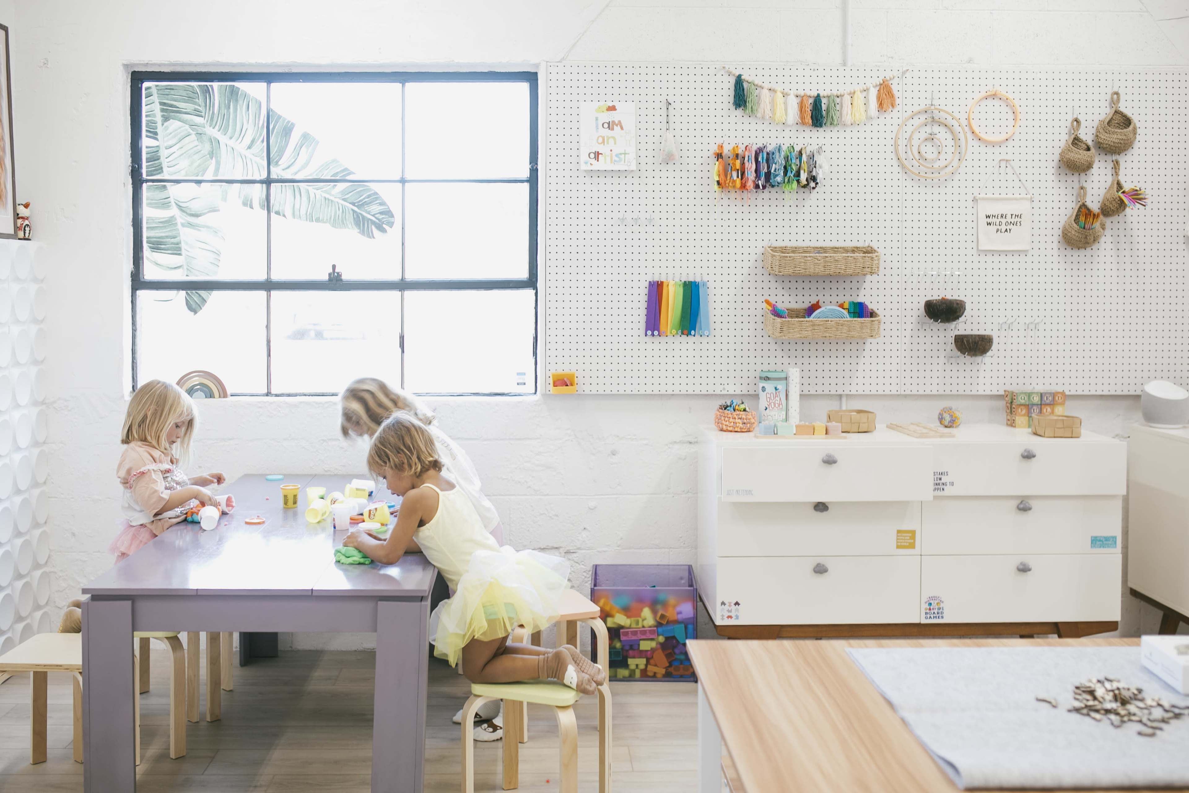 Three children are engaged in crafts at a table in a brightly lit room, with various art supplies displayed on a pegboard and a window letting in natural light.
