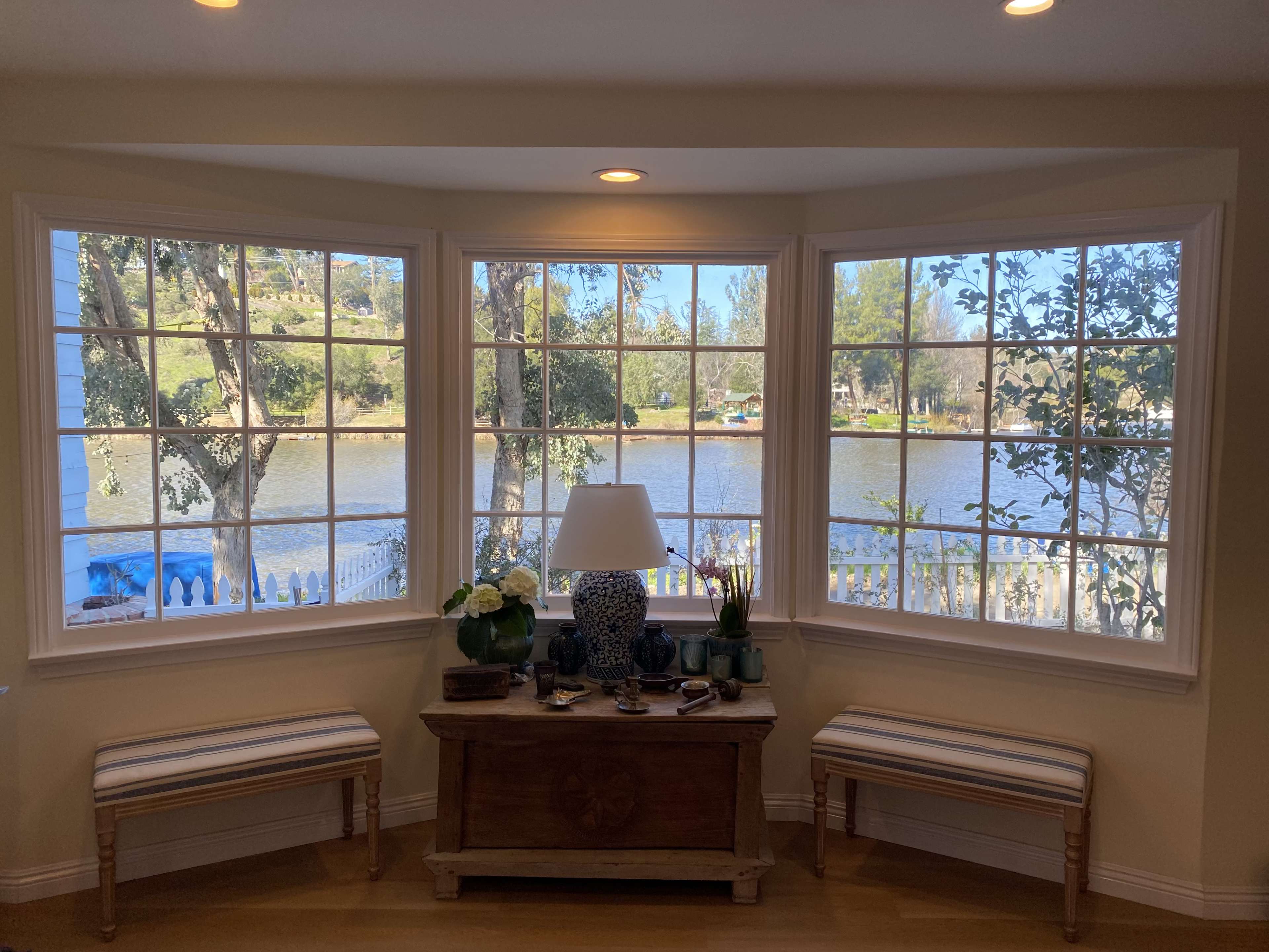 A cozy nook features two benches and a decorative table by large windows overlooking a serene lake surrounded by trees.