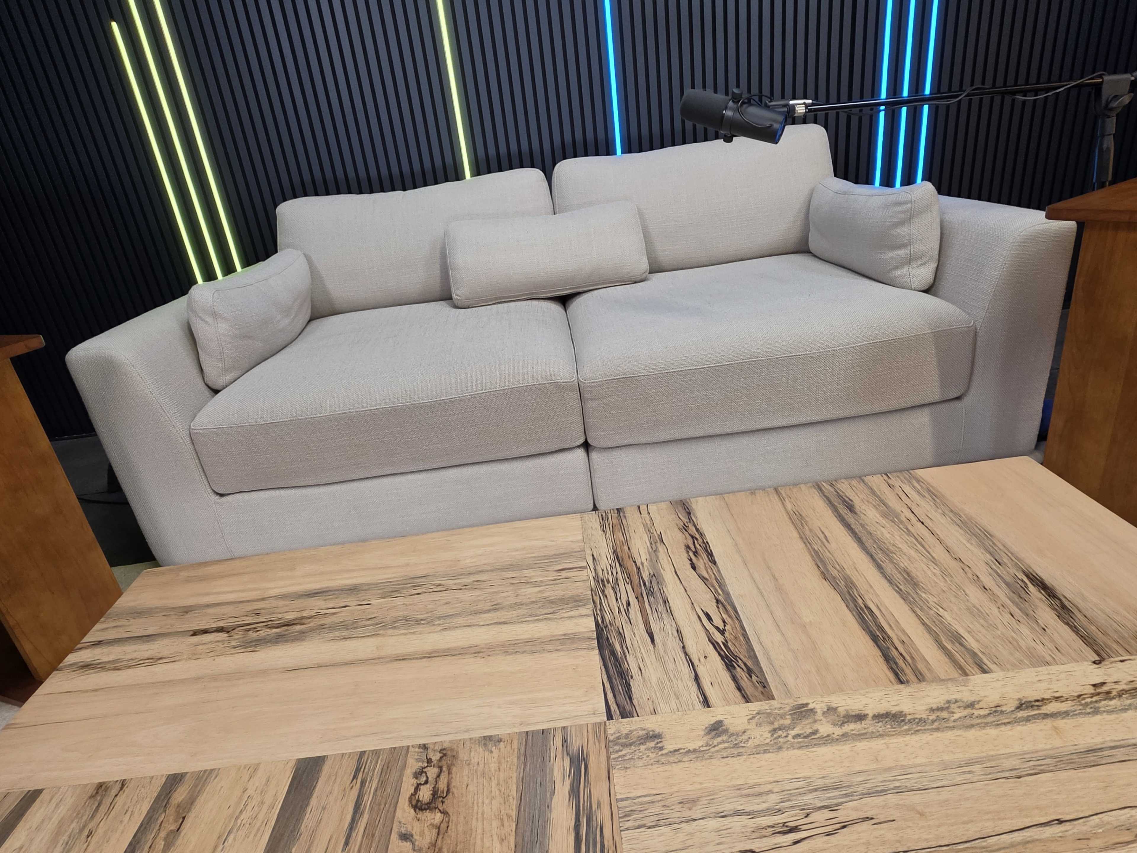 The image shows a light-colored, modern sofa placed in front of a wooden coffee table with a striped surface design, set against a backdrop of vertical black panels and colored LED lights.