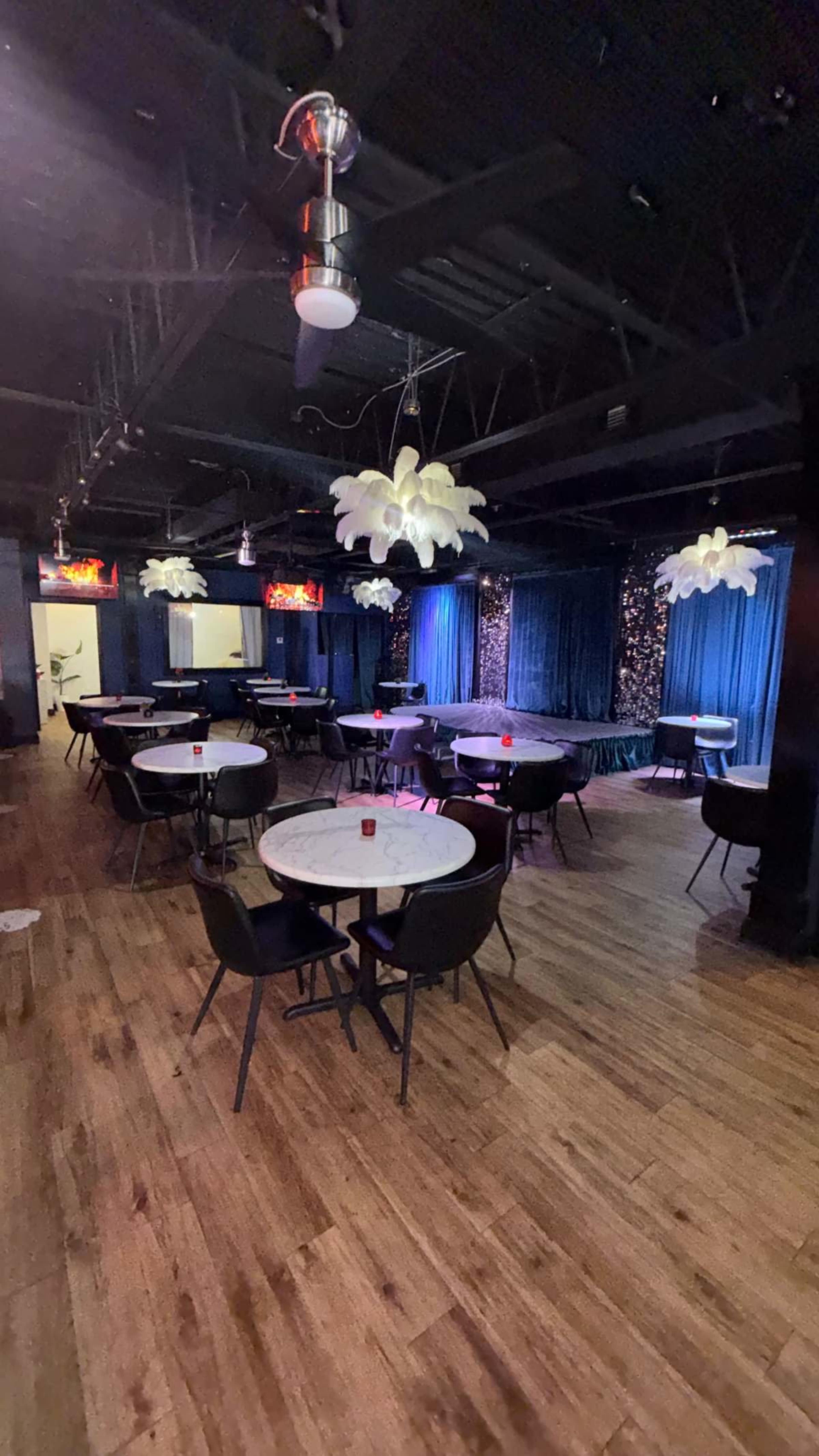 The image shows a dimly lit performance venue with round tables, chairs, and decorative light fixtures hanging from the ceiling.