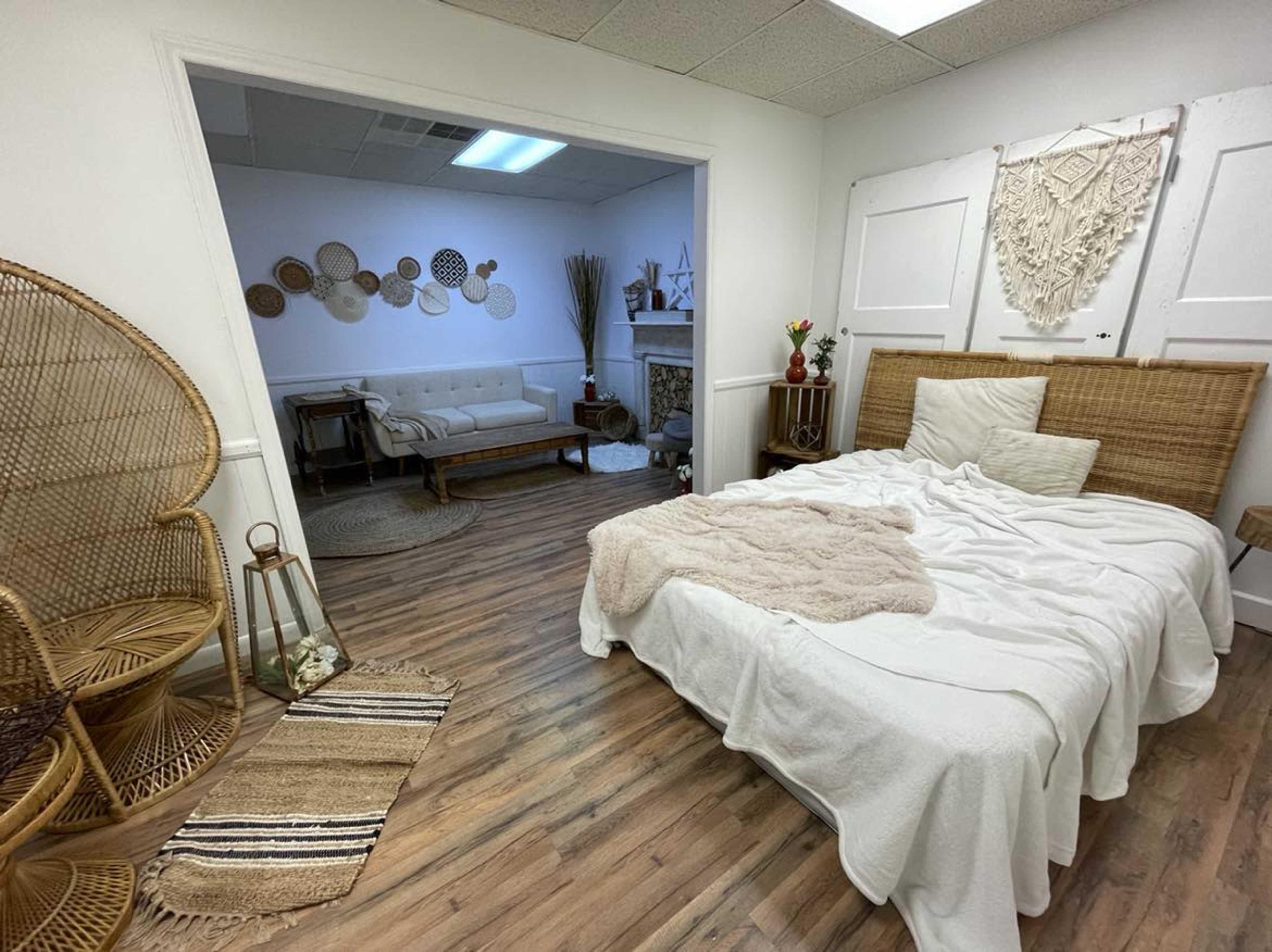 A light-colored room featuring a bed with a white blanket, a sitting area with a couch and decorative elements, and woven furniture in a cozy layout.