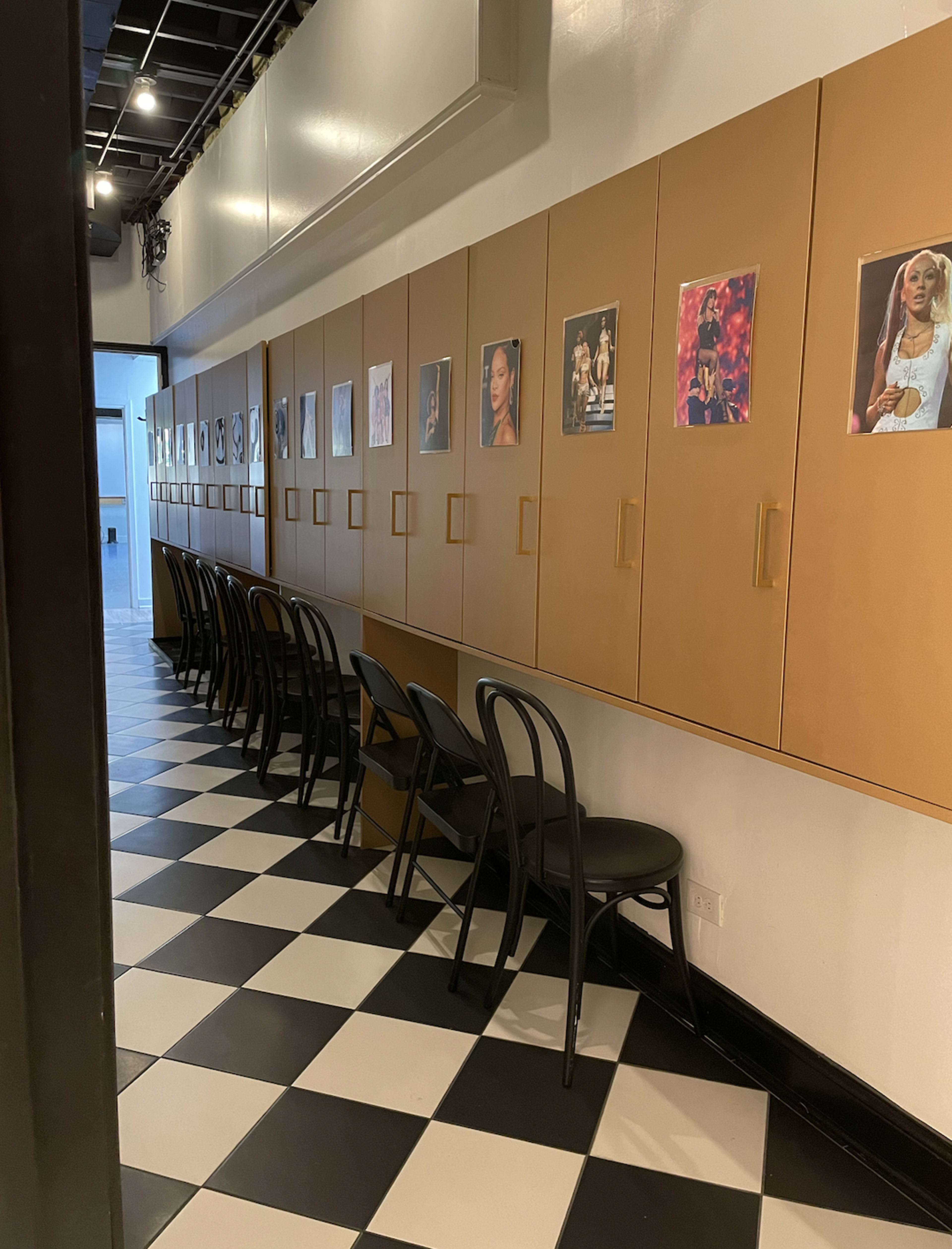 A hallway with black and white checkered flooring, lined with a row of chairs and cabinets featuring posters on the walls.