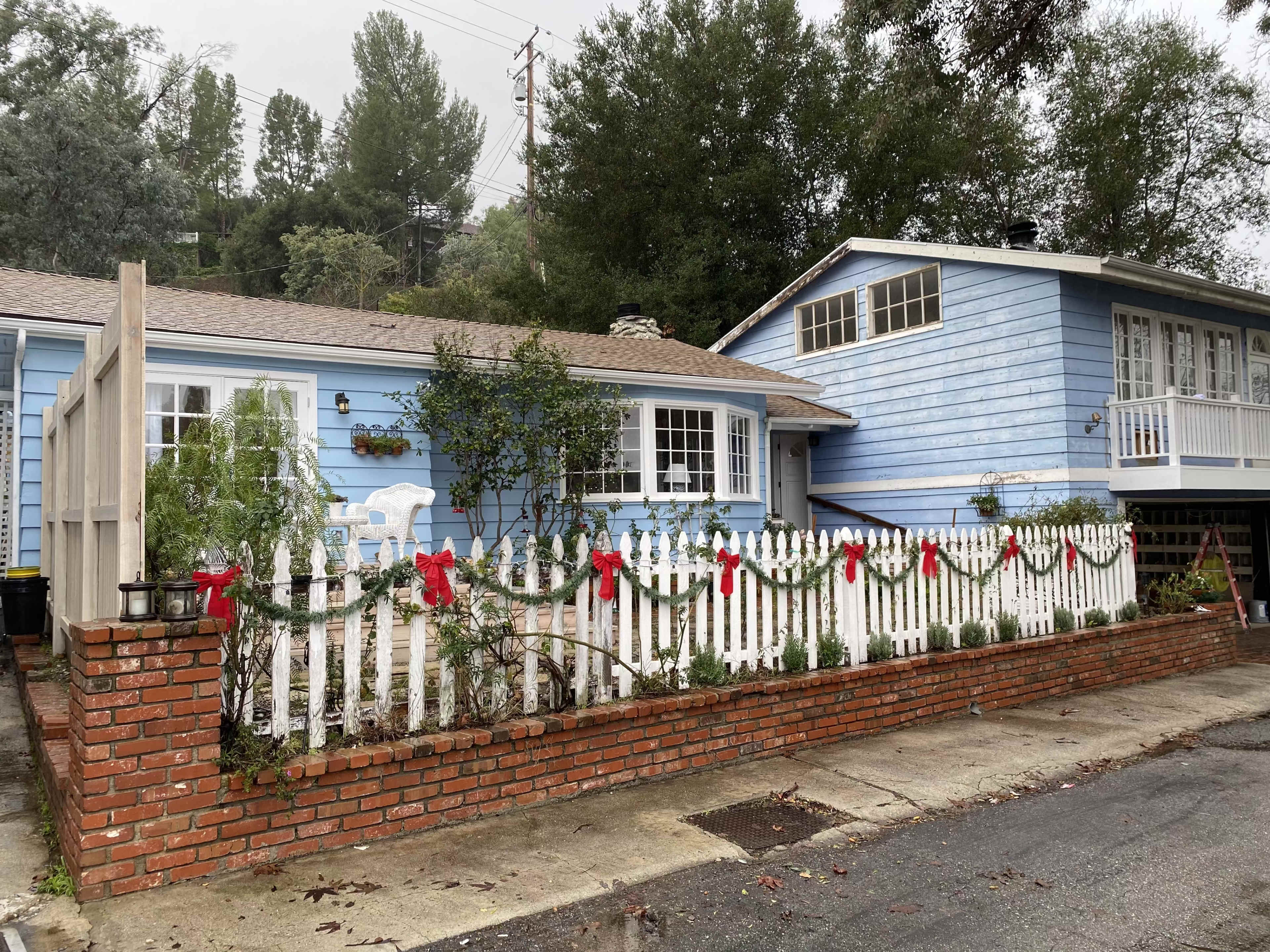 The image shows a blue house with a front yard enclosed by a white picket fence adorned with holiday garlands and bows.