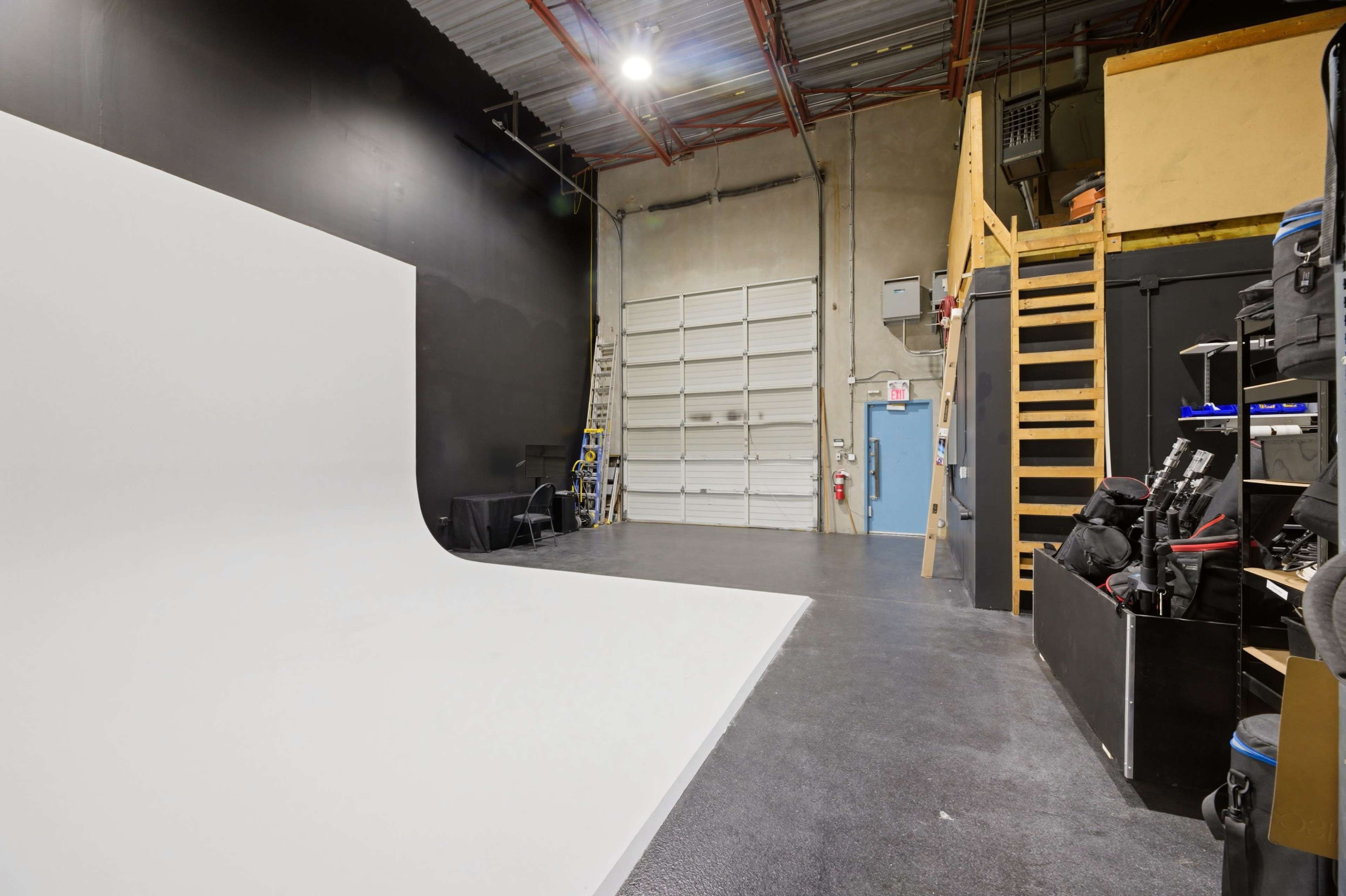 The image shows a photography studio with a large white backdrop, a black wall, a staircase leading to an upper area, and various equipment stored on the side.