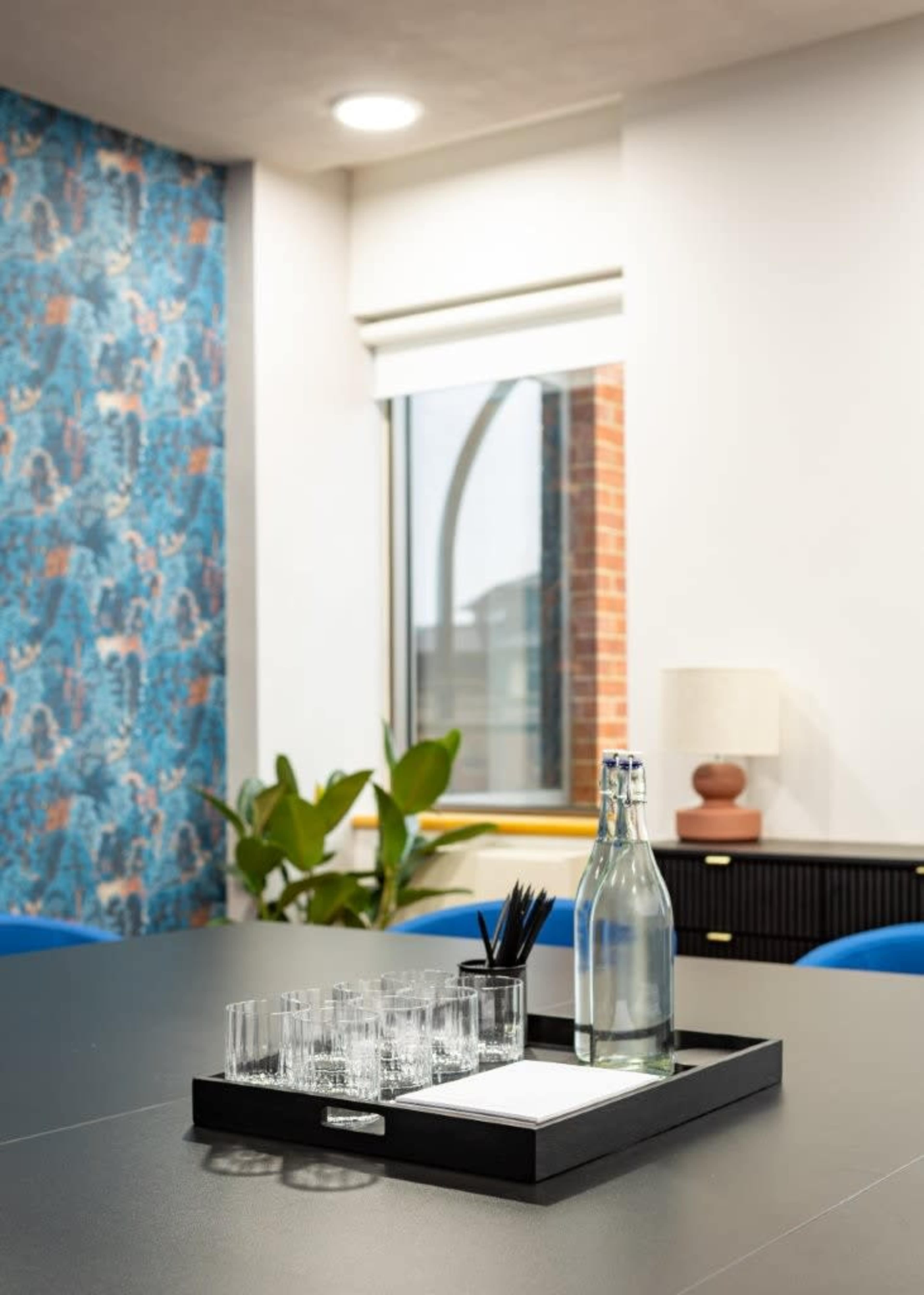 A glass tray with a bottle of water, several glasses, and a notepad is placed on a table in a bright office with a patterned wall and a window.