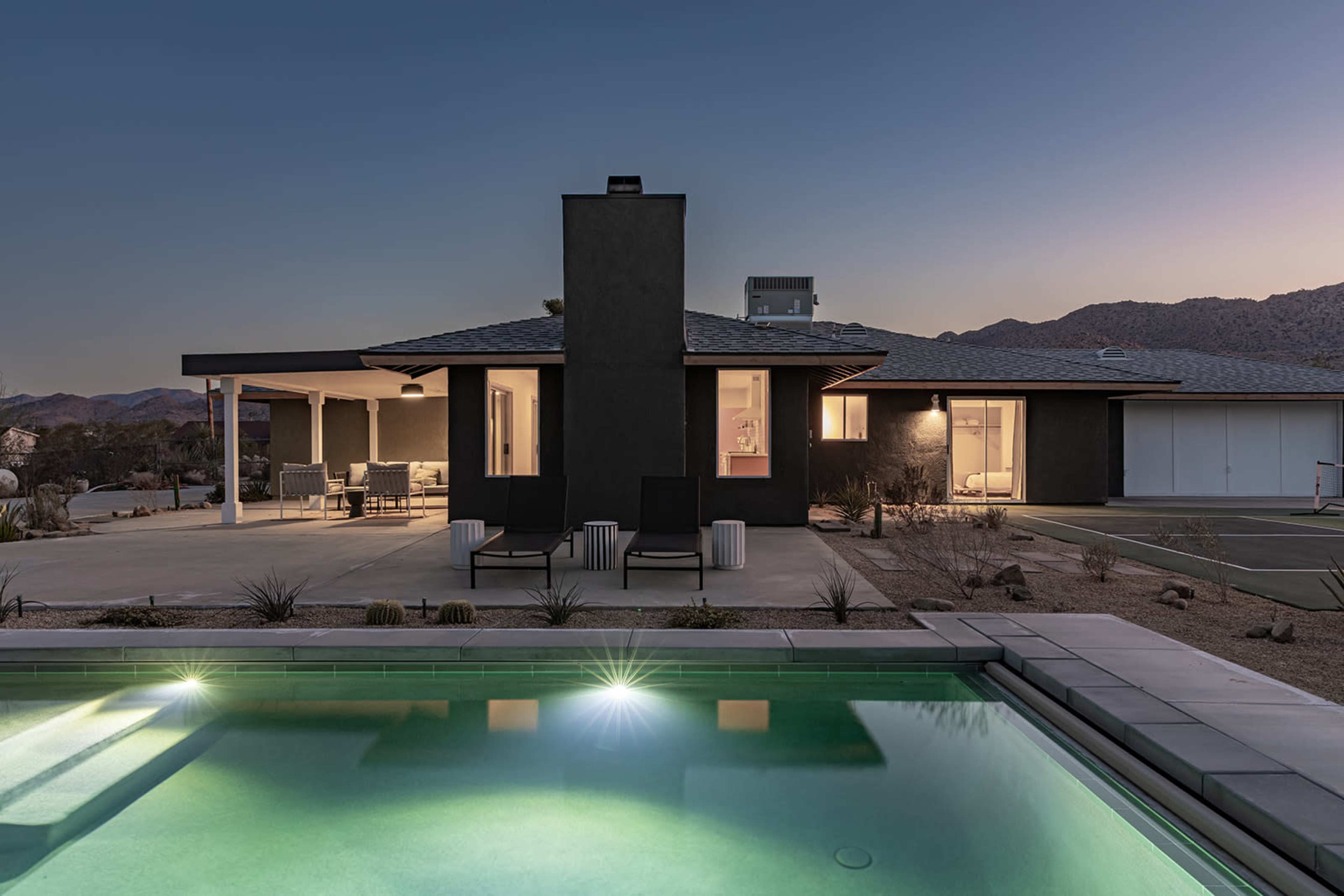A modern home with a pool is situated in a desert landscape, illuminated by soft lighting at dusk.