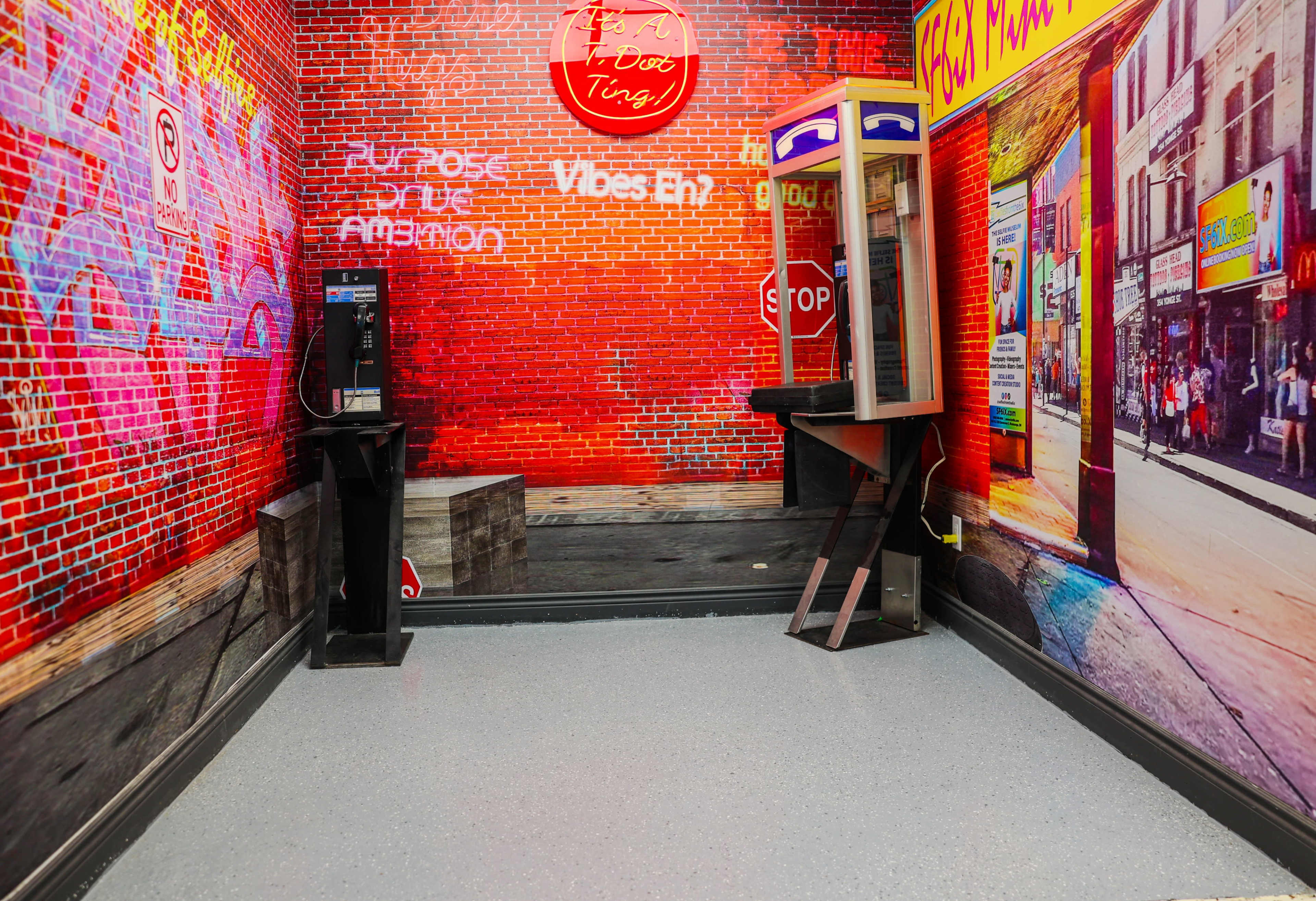 The image shows a corner of a room with a vibrant brick wall mural, featuring a payphone and a small wooden booth, alongside a gray floor with speckled finish.