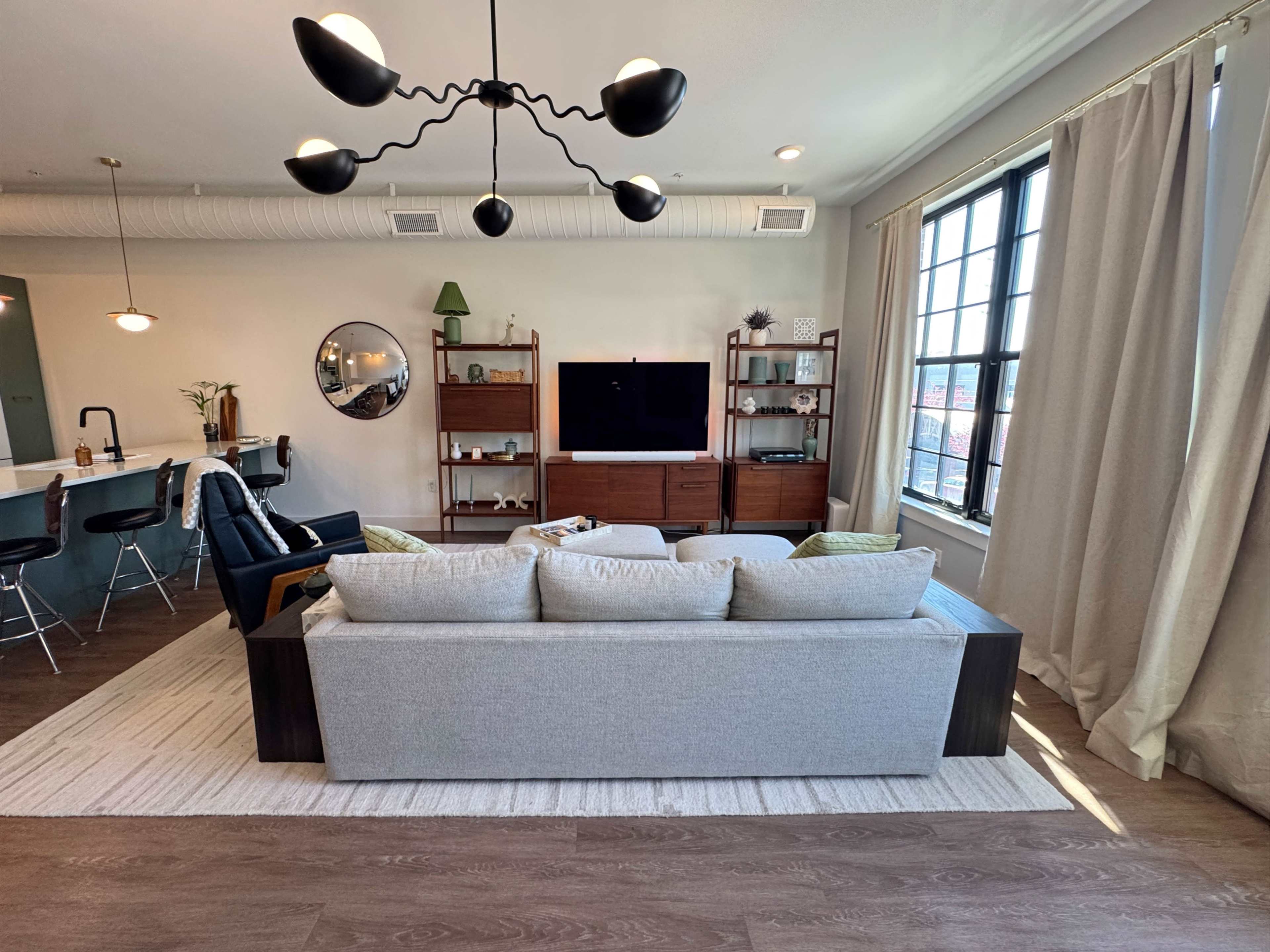 The image depicts a modern living room featuring a light gray sofa, a black accent chair, a wooden TV stand, and large windows with light beige curtains.