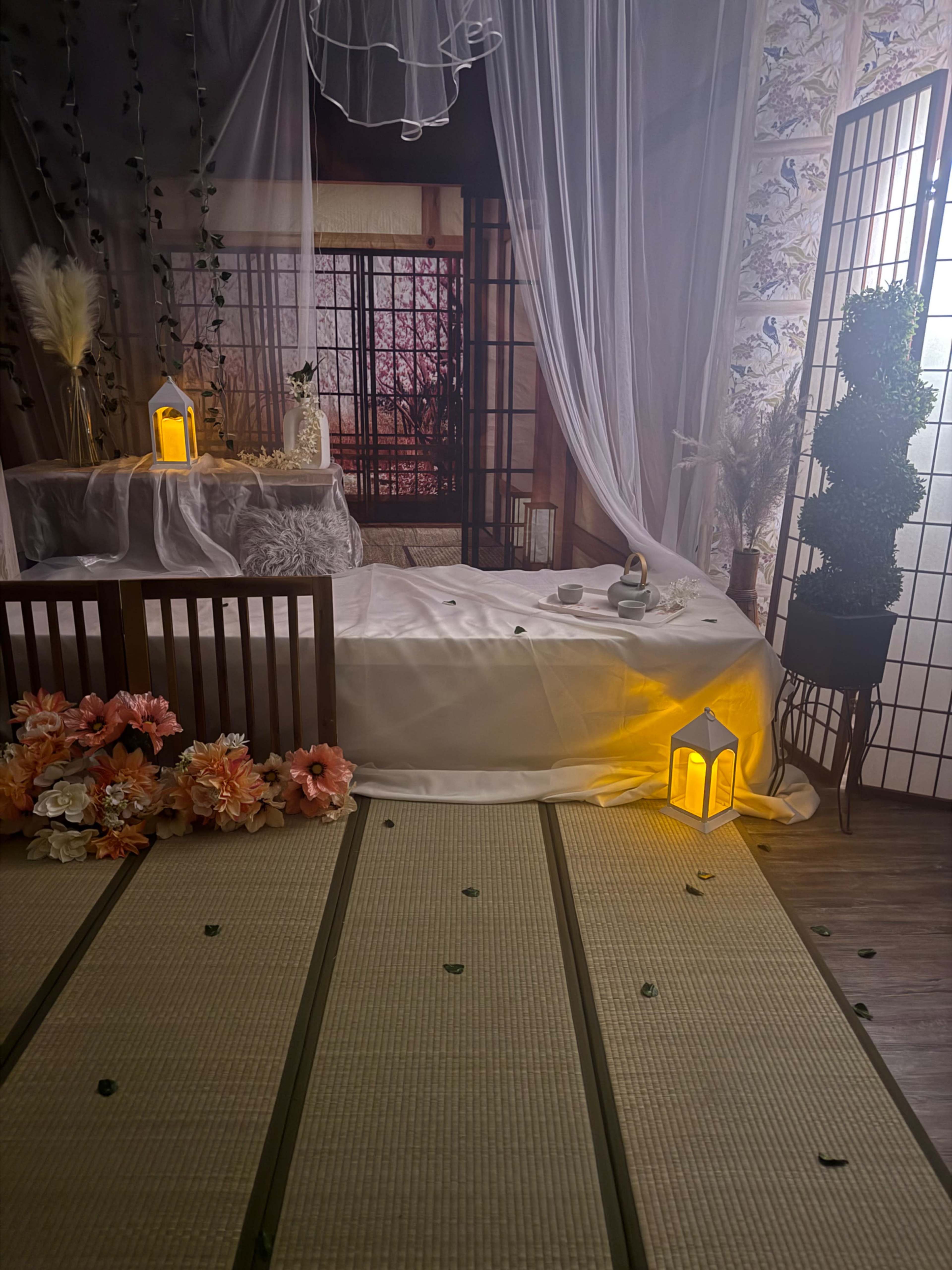 The image displays a tranquil indoor scene featuring a low wooden table surrounded by soft drapes, lanterns, and floral arrangements on a tatami mat.