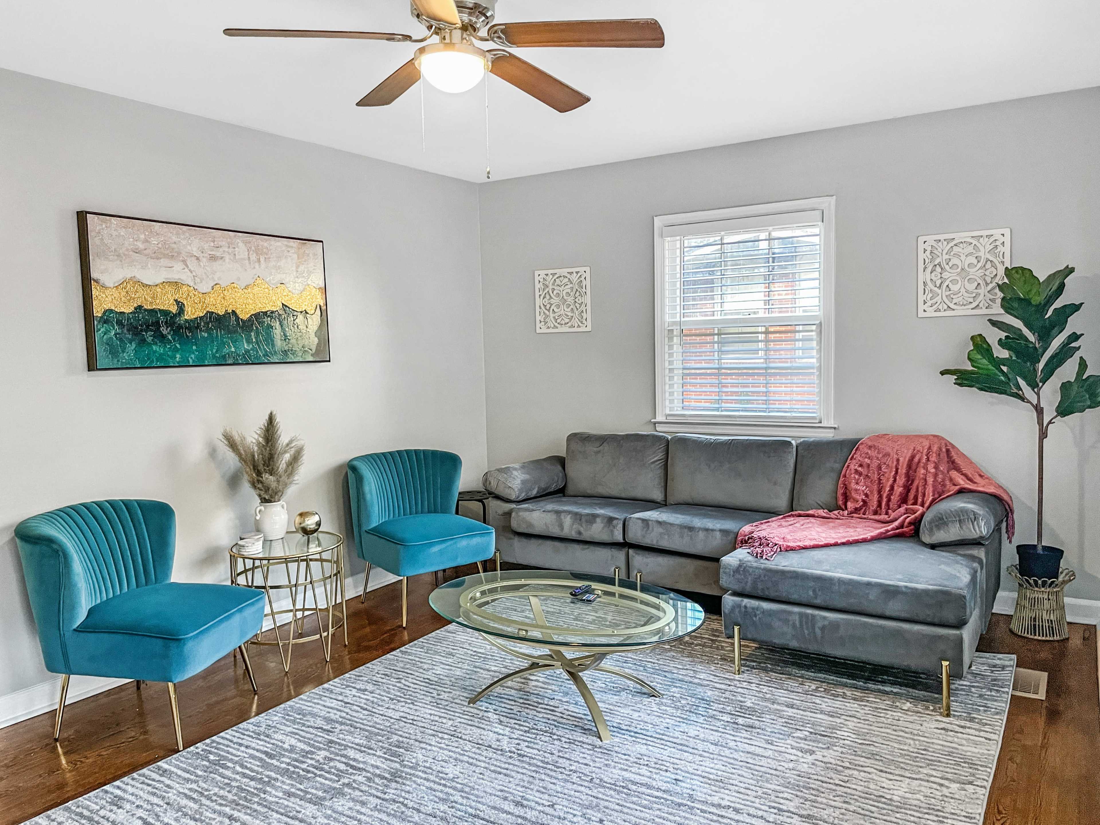 A modern living room features a gray sectional sofa, two teal chairs, a circular glass coffee table, and decorative wall art.