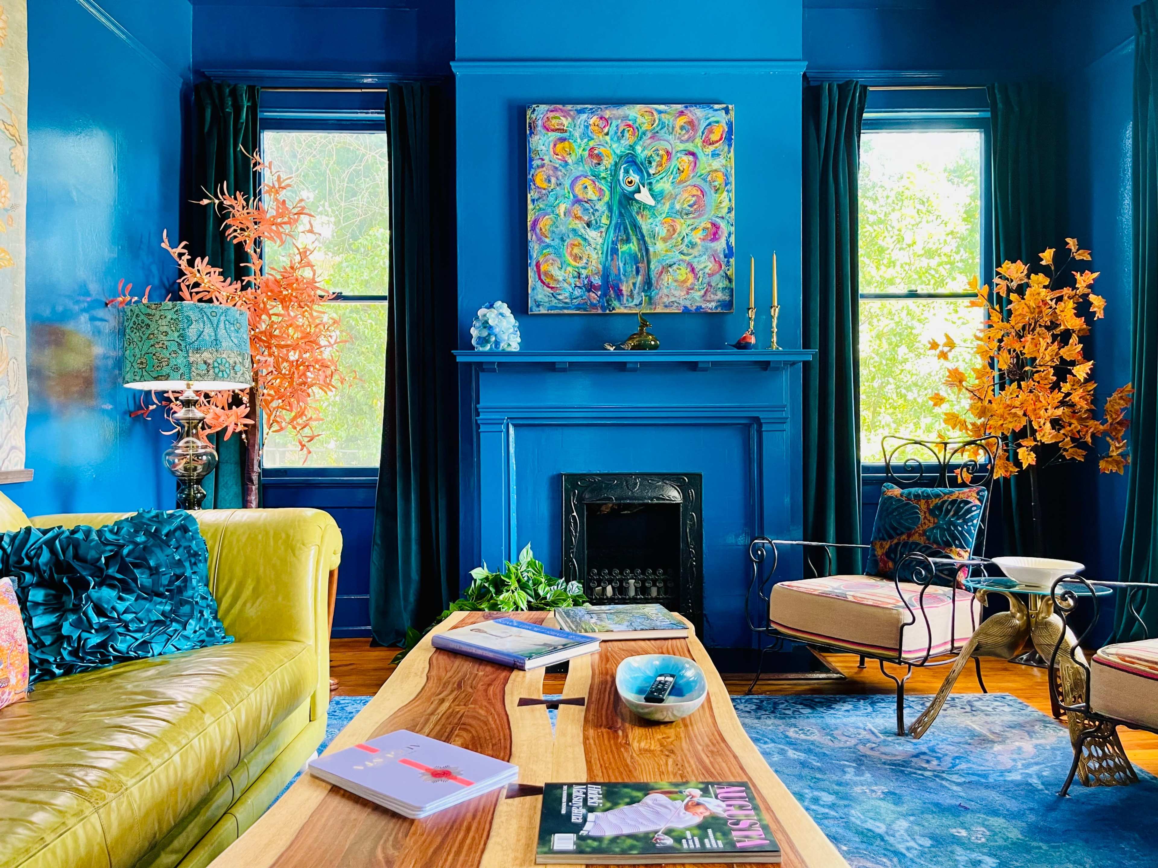The room features a vibrant blue color scheme with a yellow sofa, a wooden coffee table, and decorative plants, along with a colorful peacock painting above the fireplace.