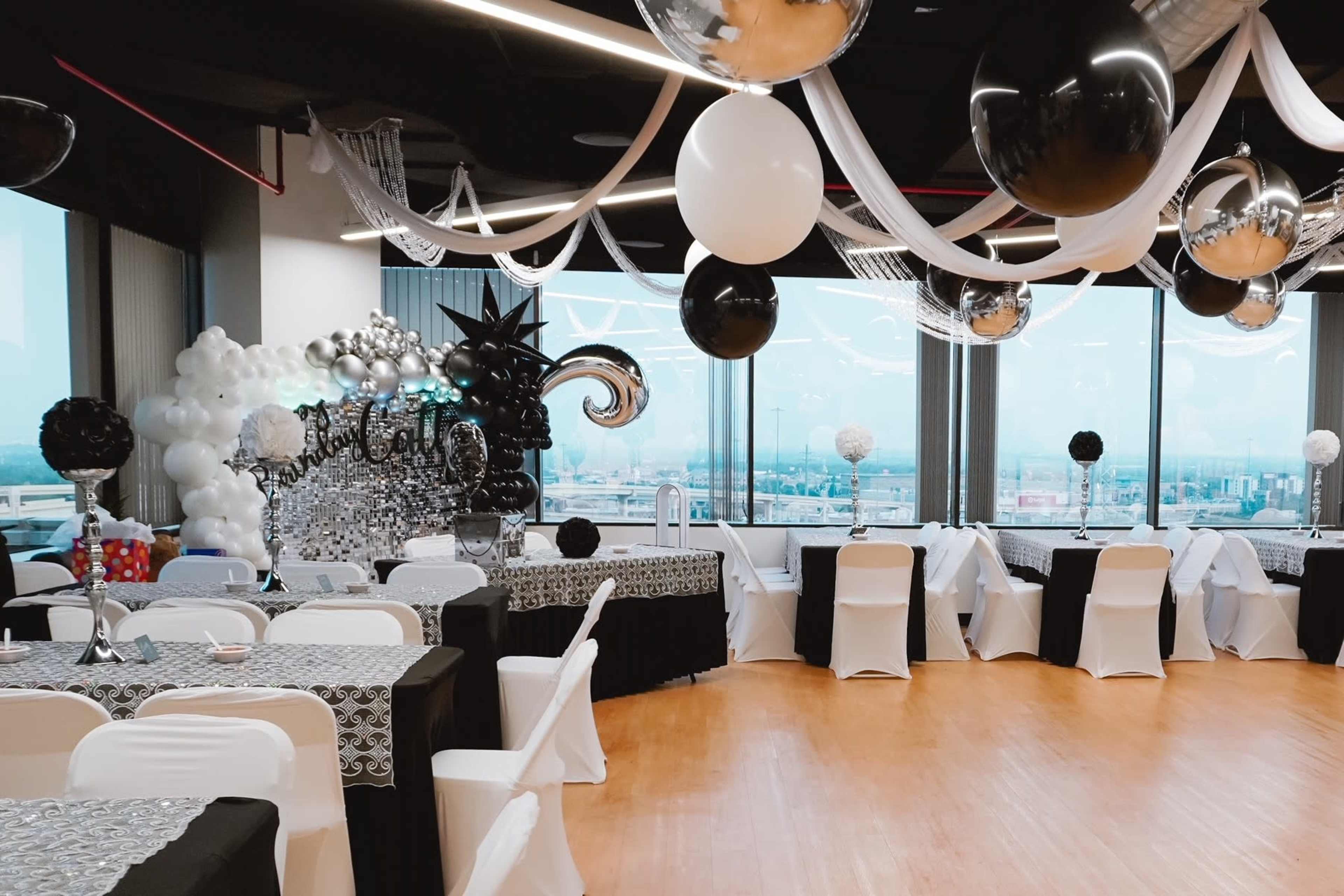 The event space features black and white decorations, with tables set and balloons hanging from the ceiling overlooking a city view.
