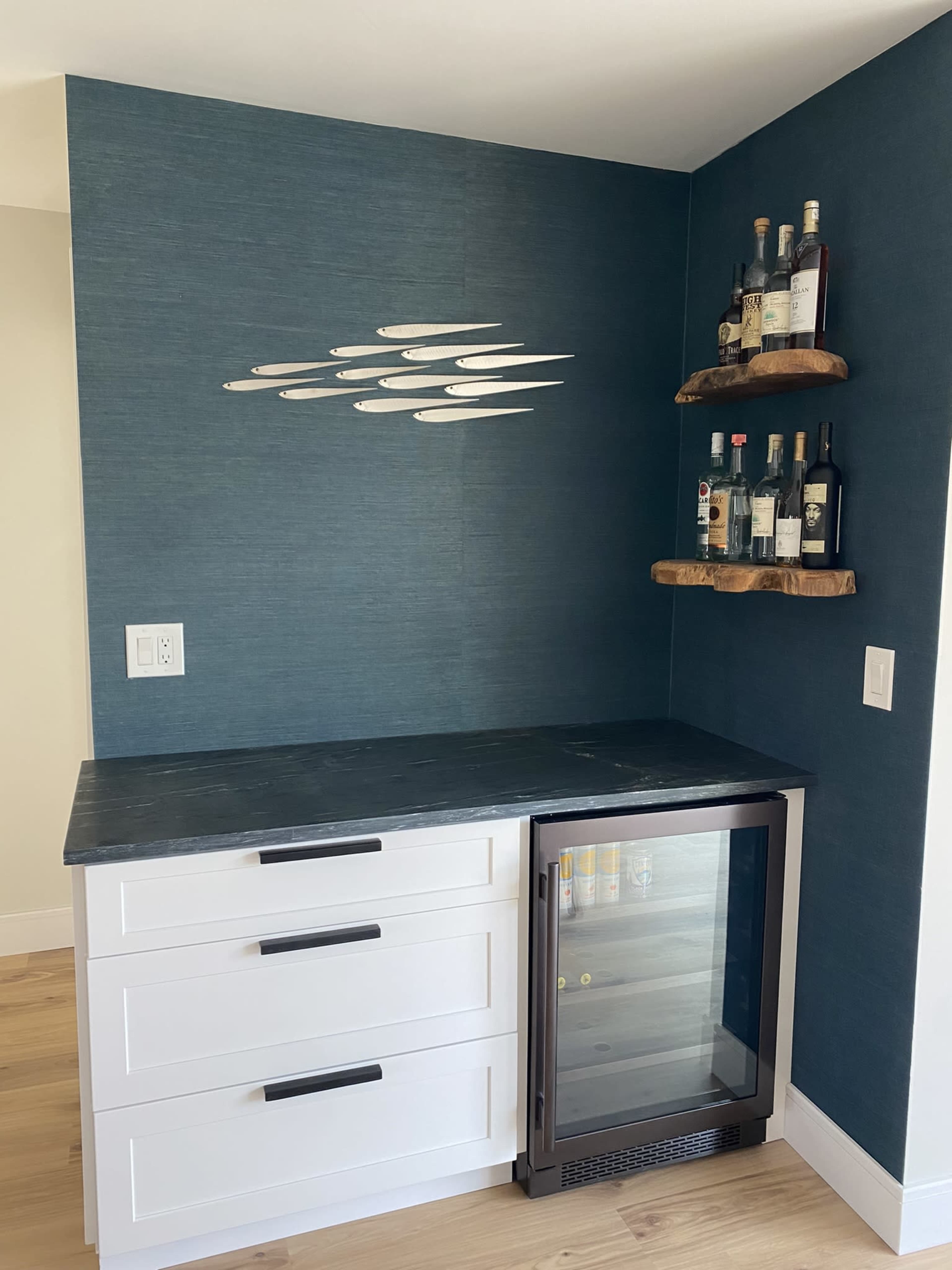 The image shows a corner bar area featuring a blue textured wall, a white cabinetry unit with three drawers, a wine cooler, and two wooden shelves displaying several bottles of liquor.