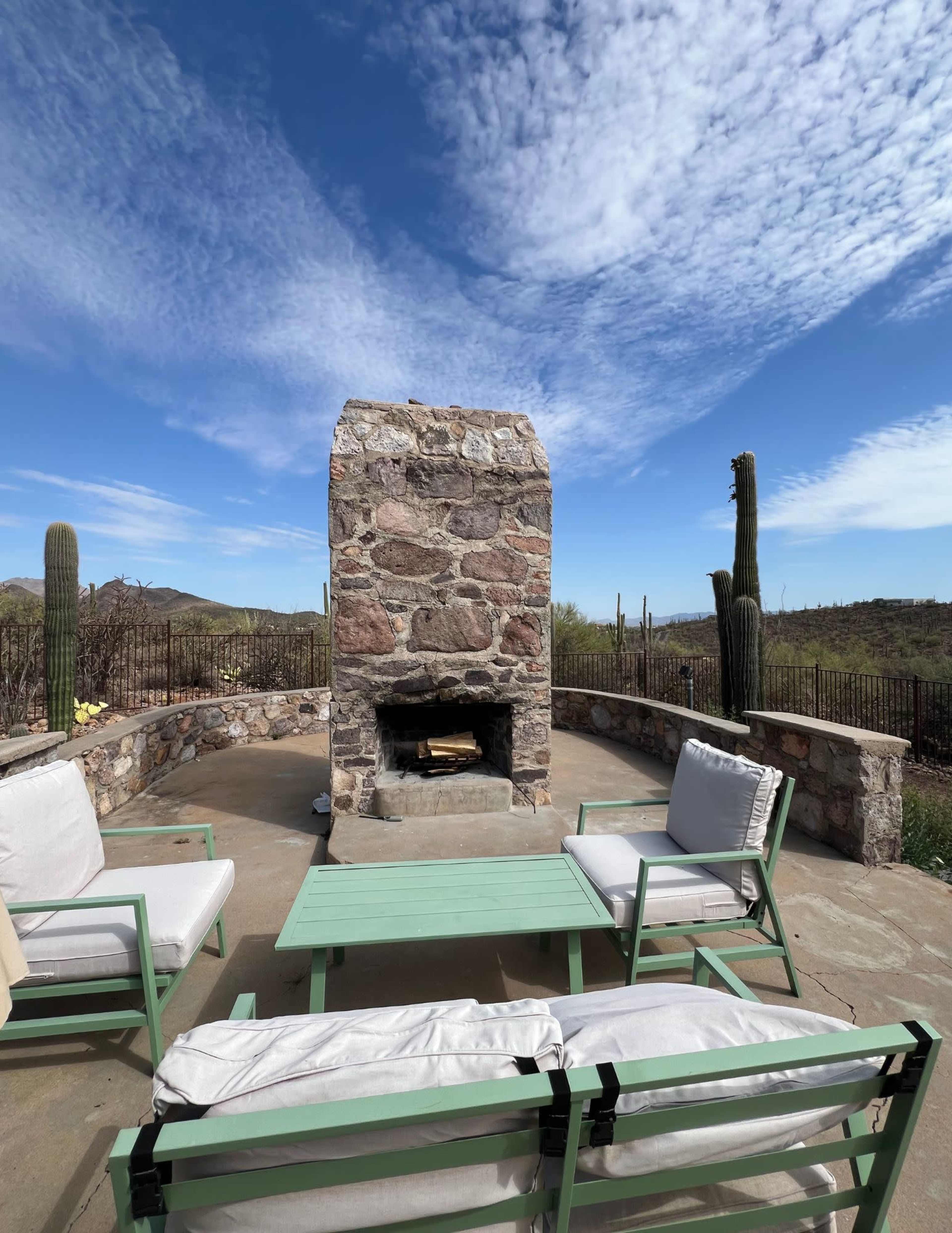 The image shows a stone fireplace surrounded by outdoor seating on a patio under a clear blue sky with sparse clouds and desert vegetation in the background.