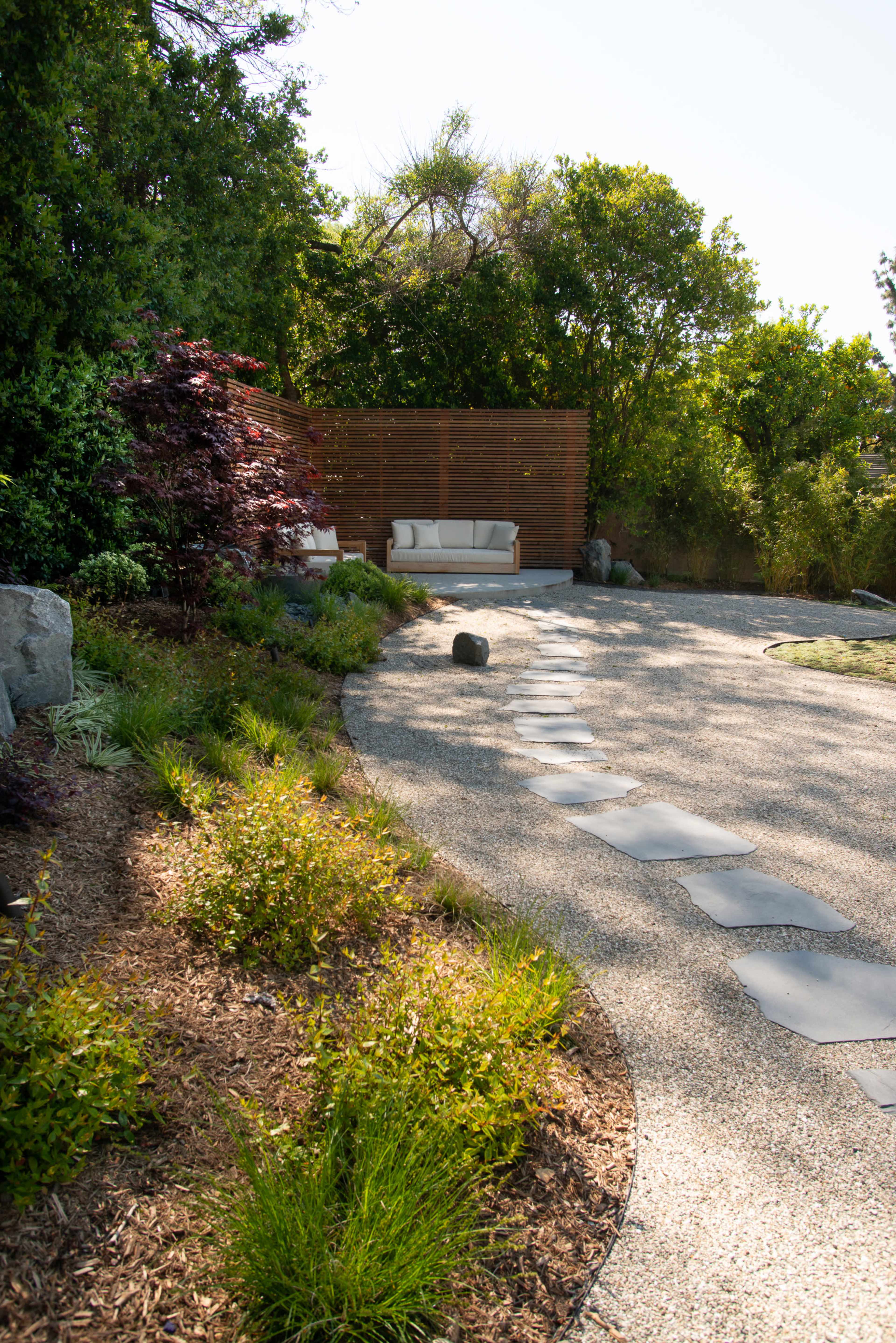 A curved stone pathway leads to a seating area surrounded by greenery and a wooden privacy screen.