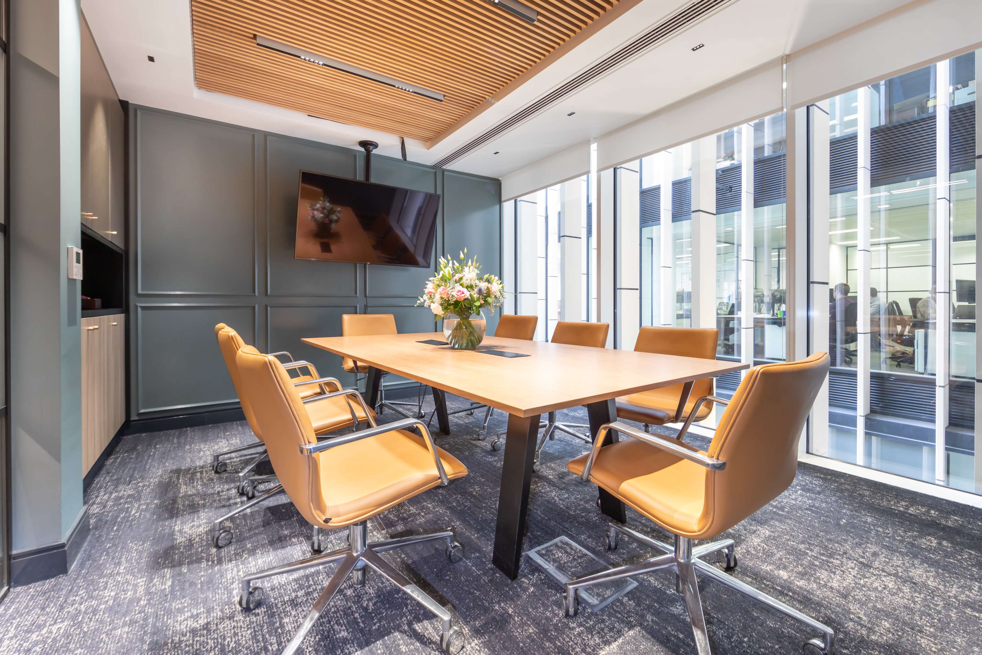 A modern conference room features a large wooden table surrounded by brown leather chairs, with a wall-mounted TV and large windows providing natural light.