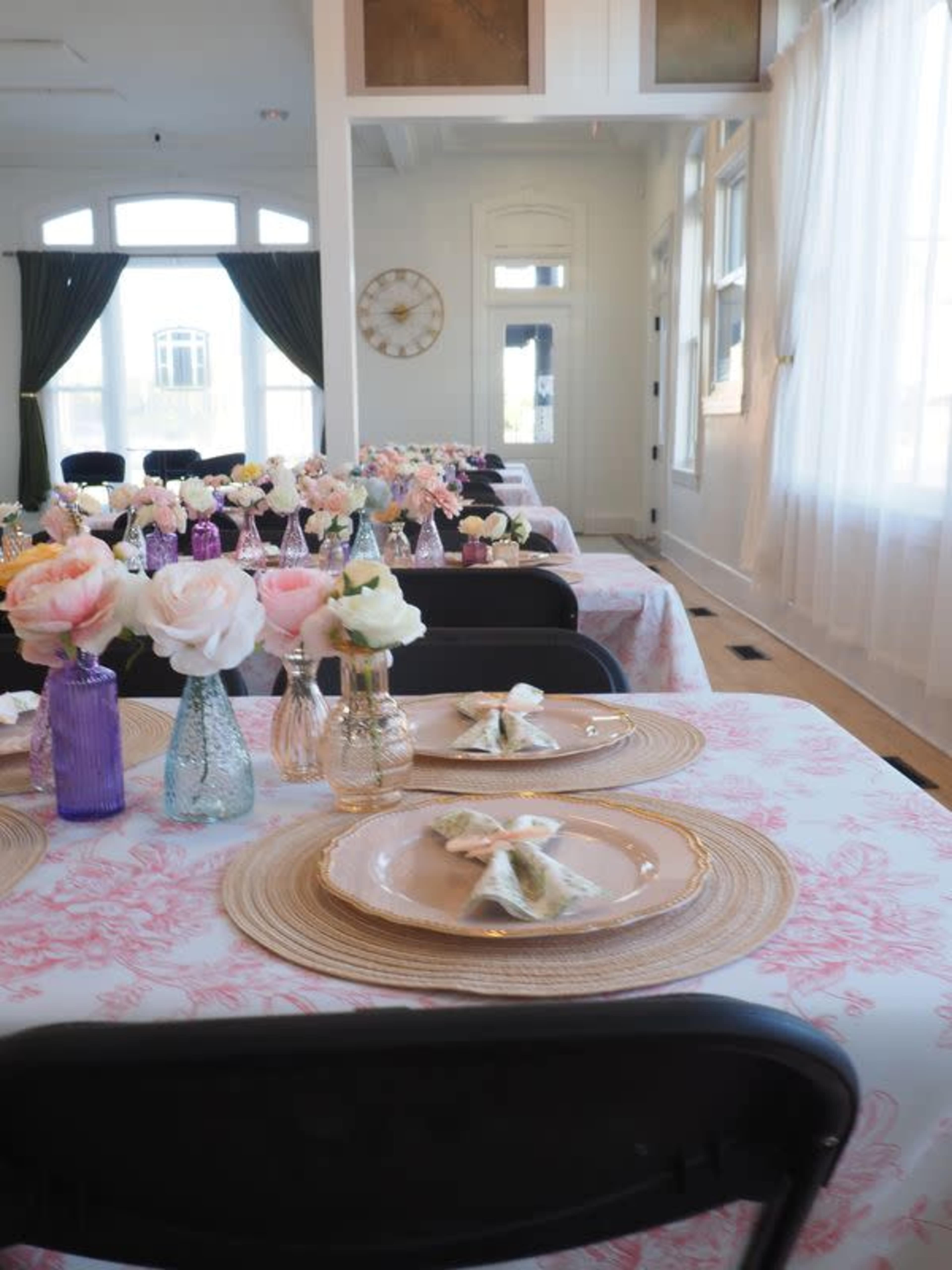 The image shows an elegantly set dining area with tables covered in pink floral tablecloths, adorned with vases of roses and decorative placemats.