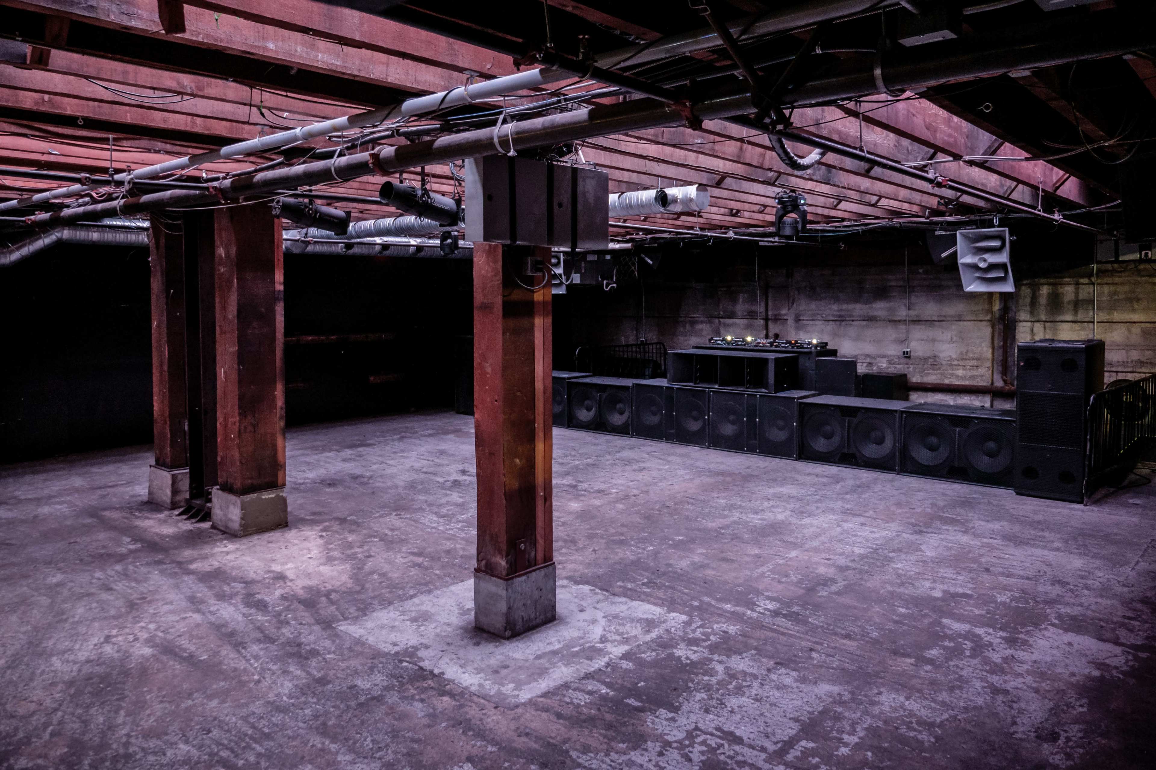 An empty underground venue with concrete walls, wooden beams, and a sound setup along one side.