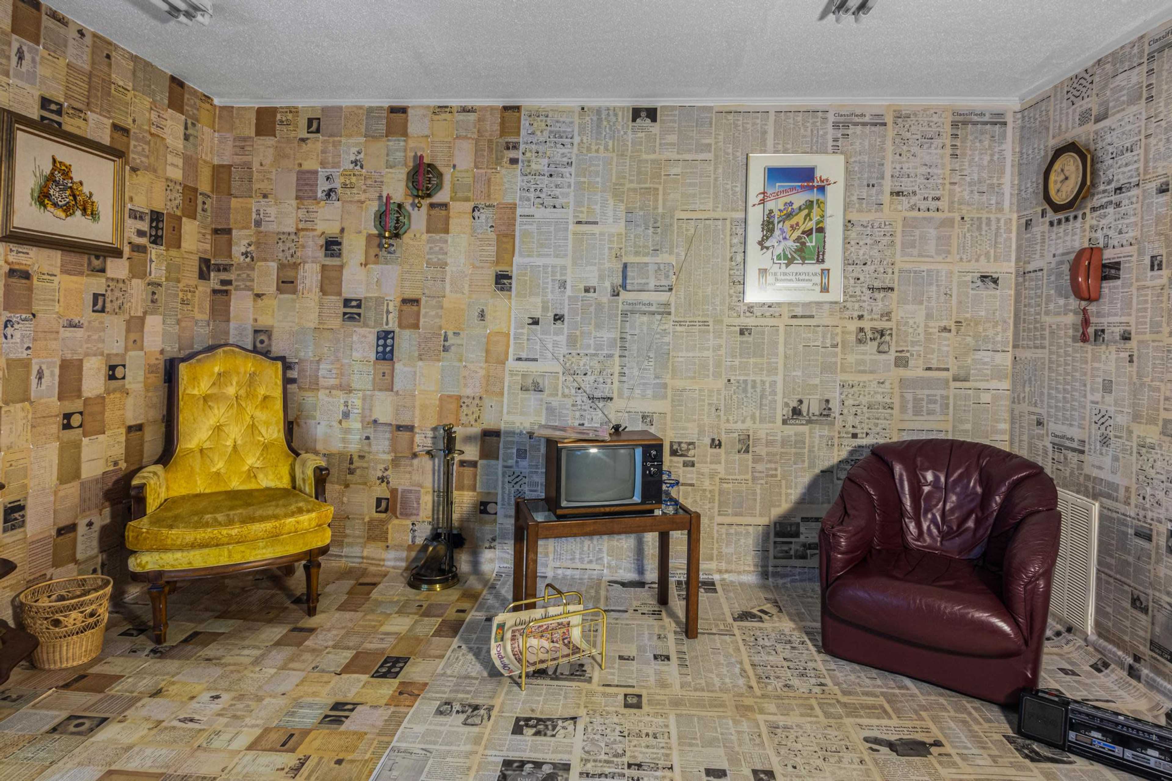 The room features walls and floors covered in newspaper clippings, with a yellow armchair, a red leather chair, a wooden table, a vintage television, a guitar, and a retro wall clock.