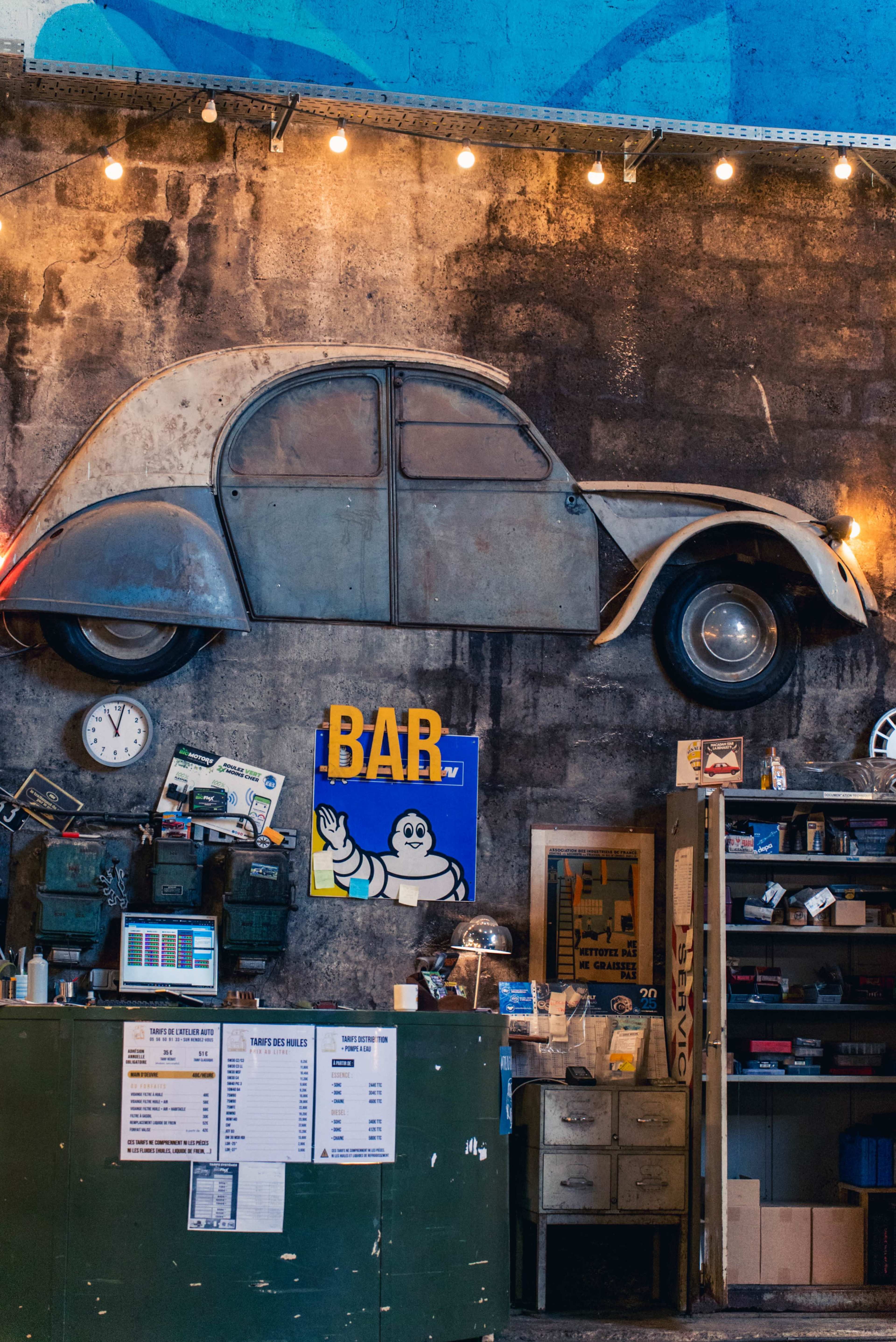 The image shows an industrial interior with a vintage car mounted on the wall, a bar sign, a clock, and various storage cabinets.