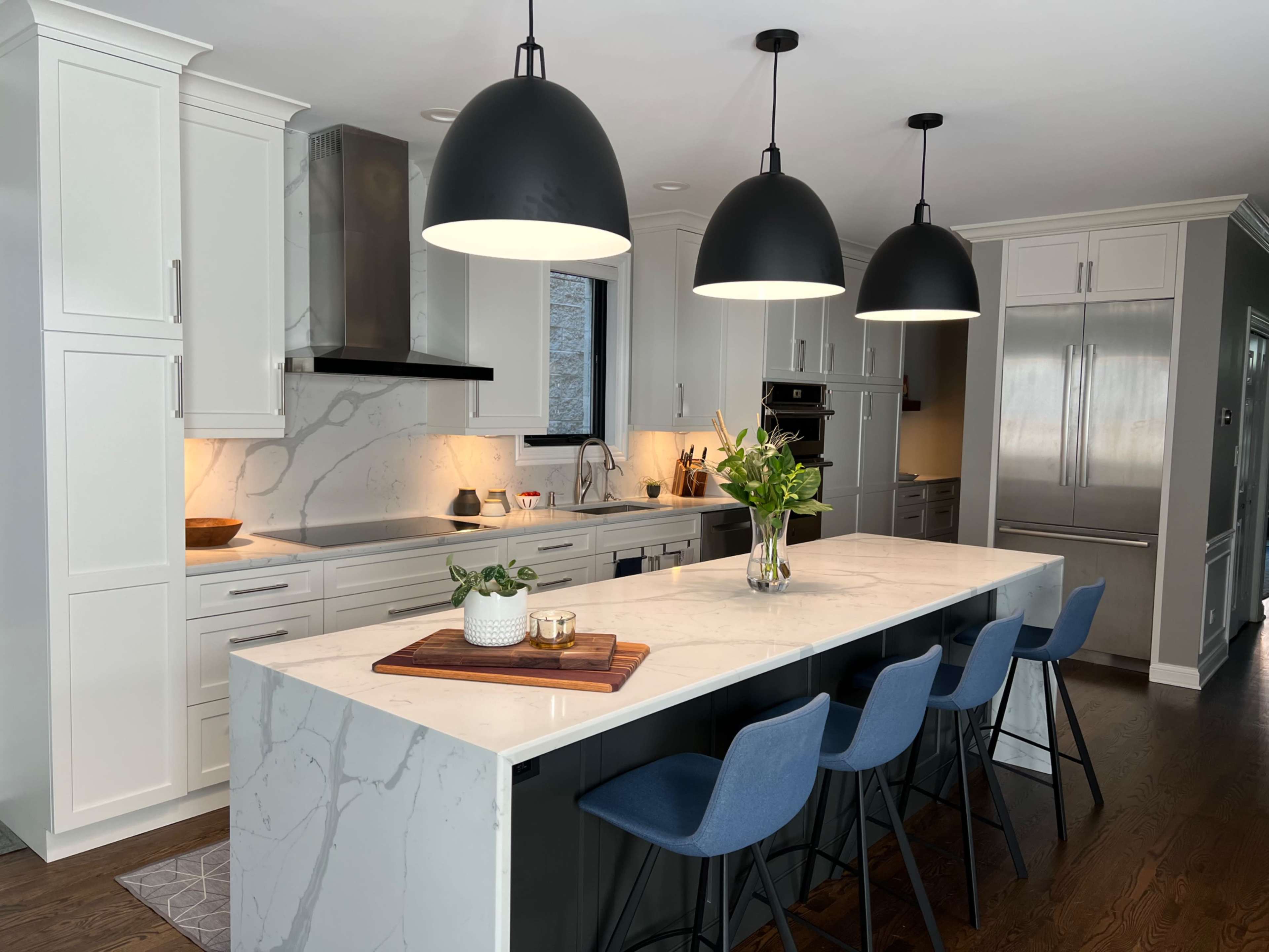 A modern kitchen features a large island with a marble countertop and blue bar stools, illuminated by three black pendant lights.