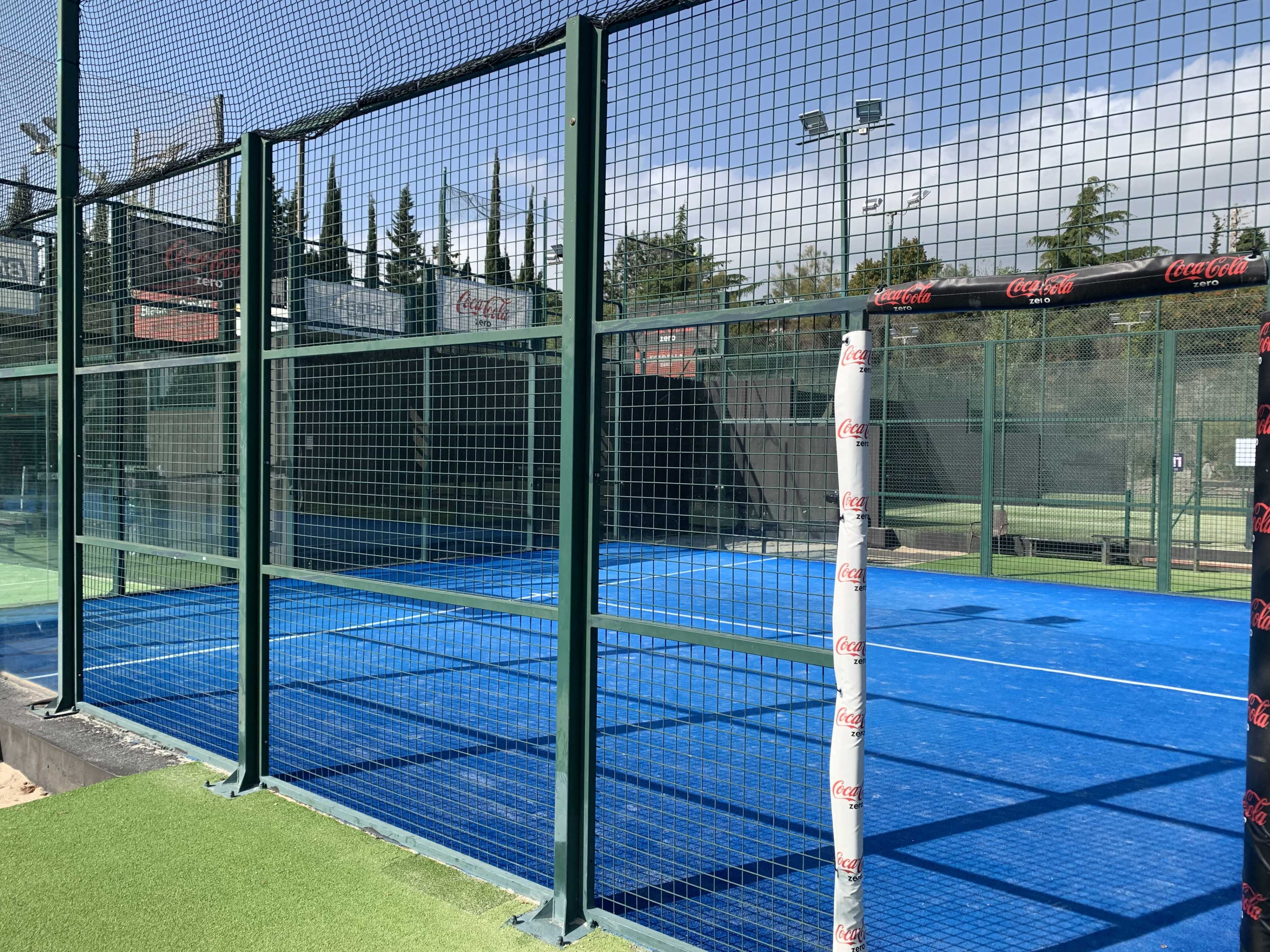 A blue padel court enclosed by green metal fencing, with a visible net and surrounding facilities.