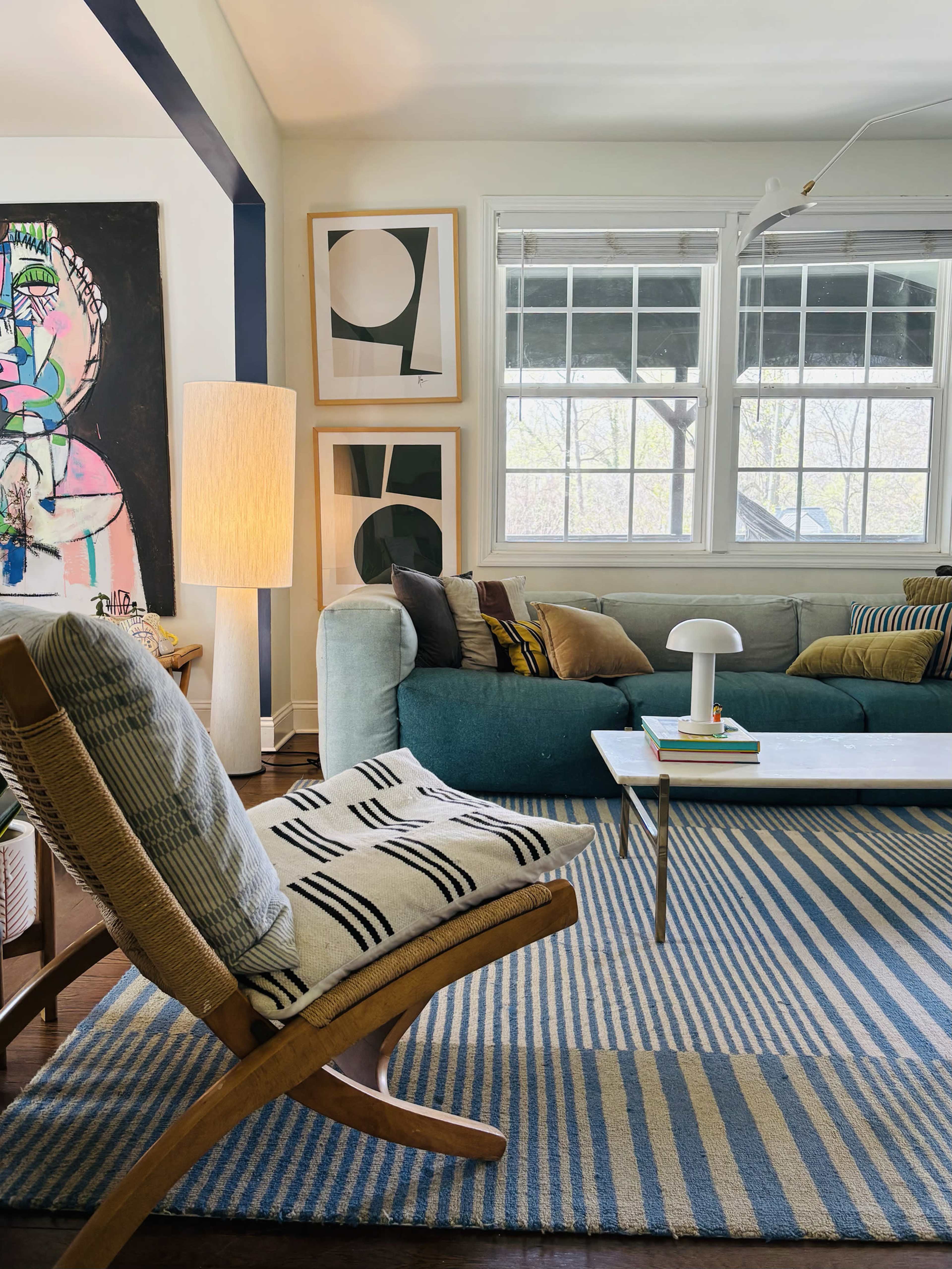 The image shows a cozy living room featuring a teal sofa, a patterned rug with blue stripes, a wooden chair, and abstract wall art.